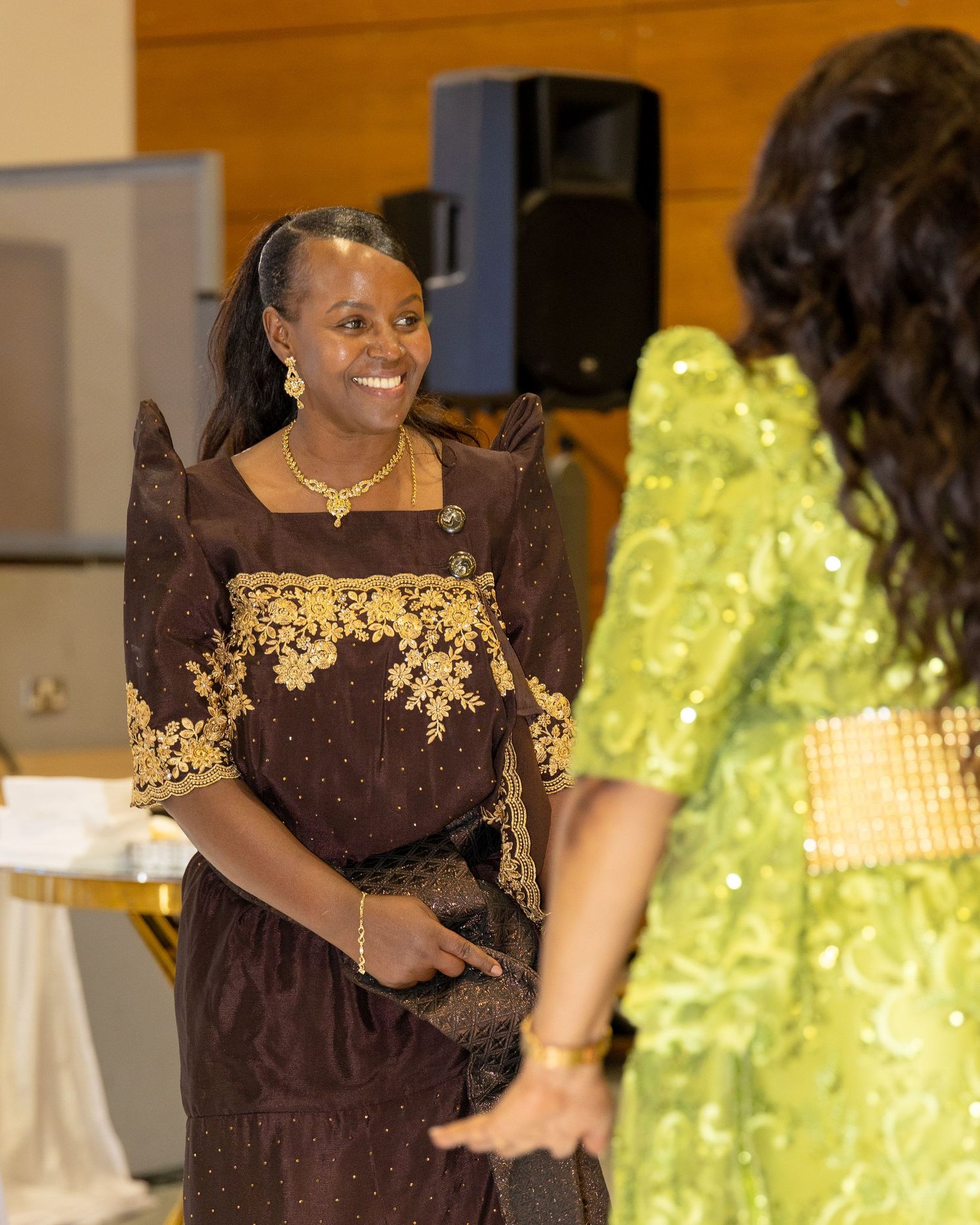 Woman in brown and gold traditional African dress smiles warmly at wedding reception event.
