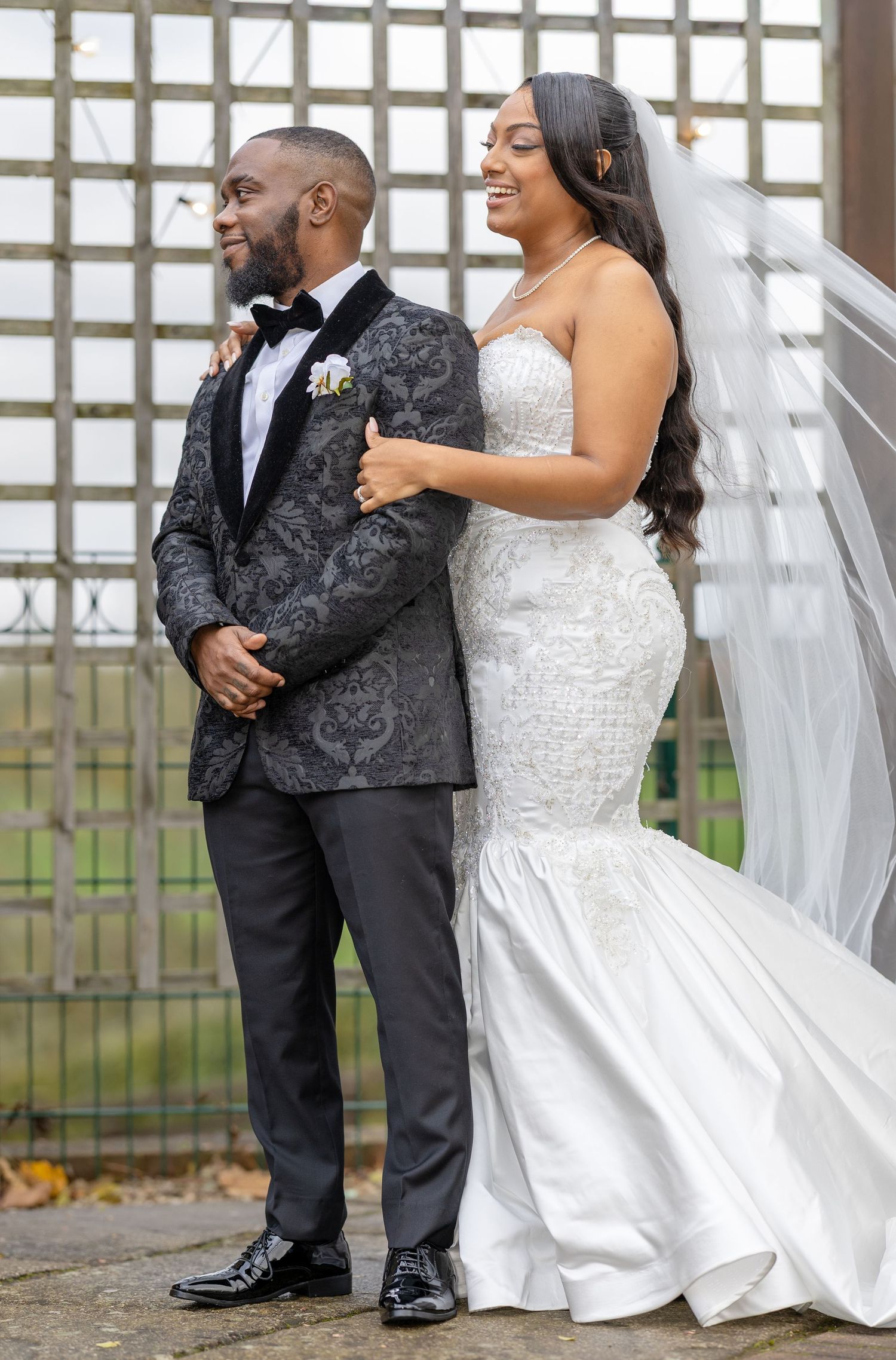 Bride and groom share a joyful moment in wedding attire, groom in patterned tuxedo, bride in beaded mermaid gown with veil.