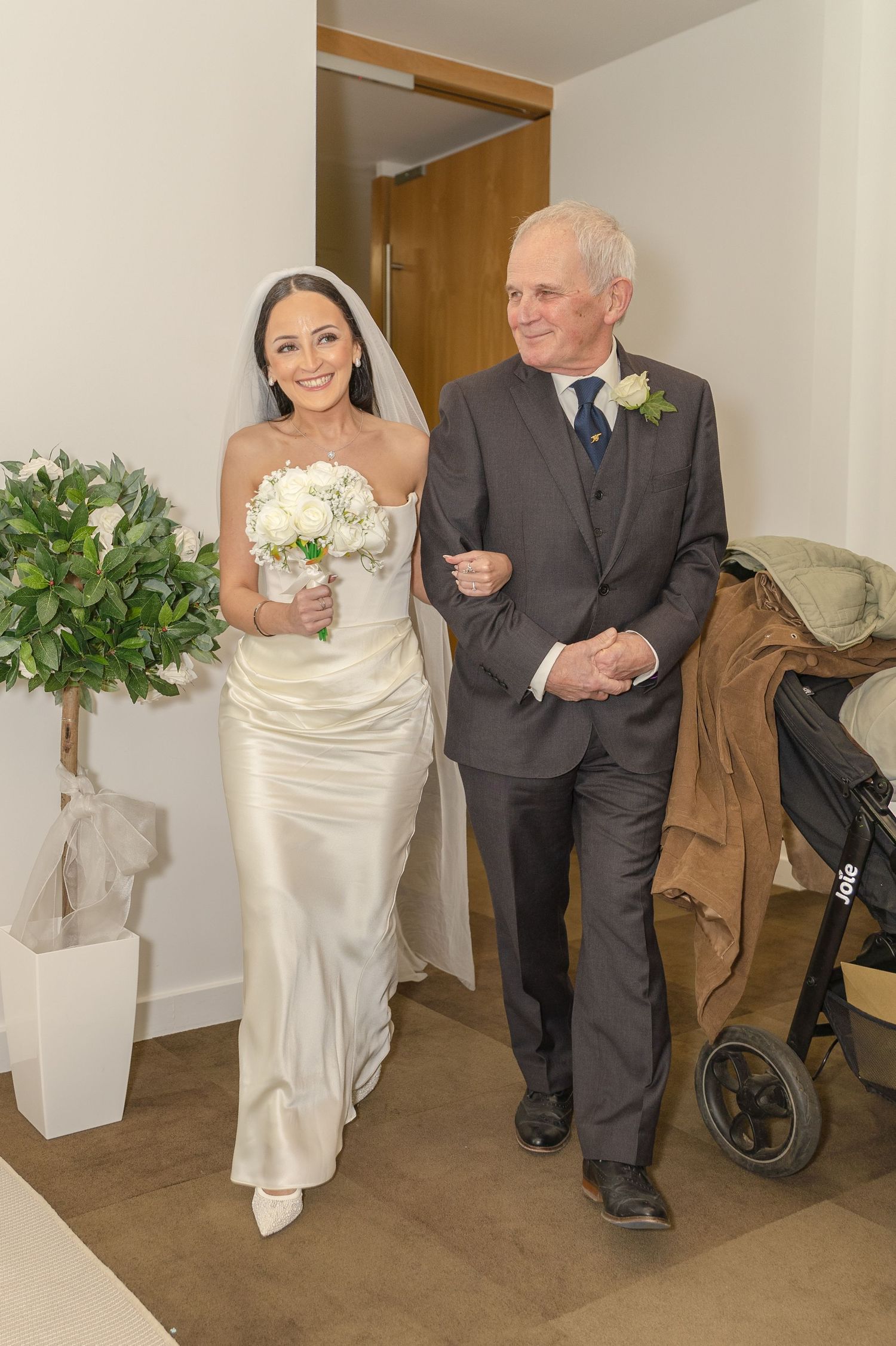 Elegant bride holding white bouquet walks beside older man in grey suit through a modern wedding venue.