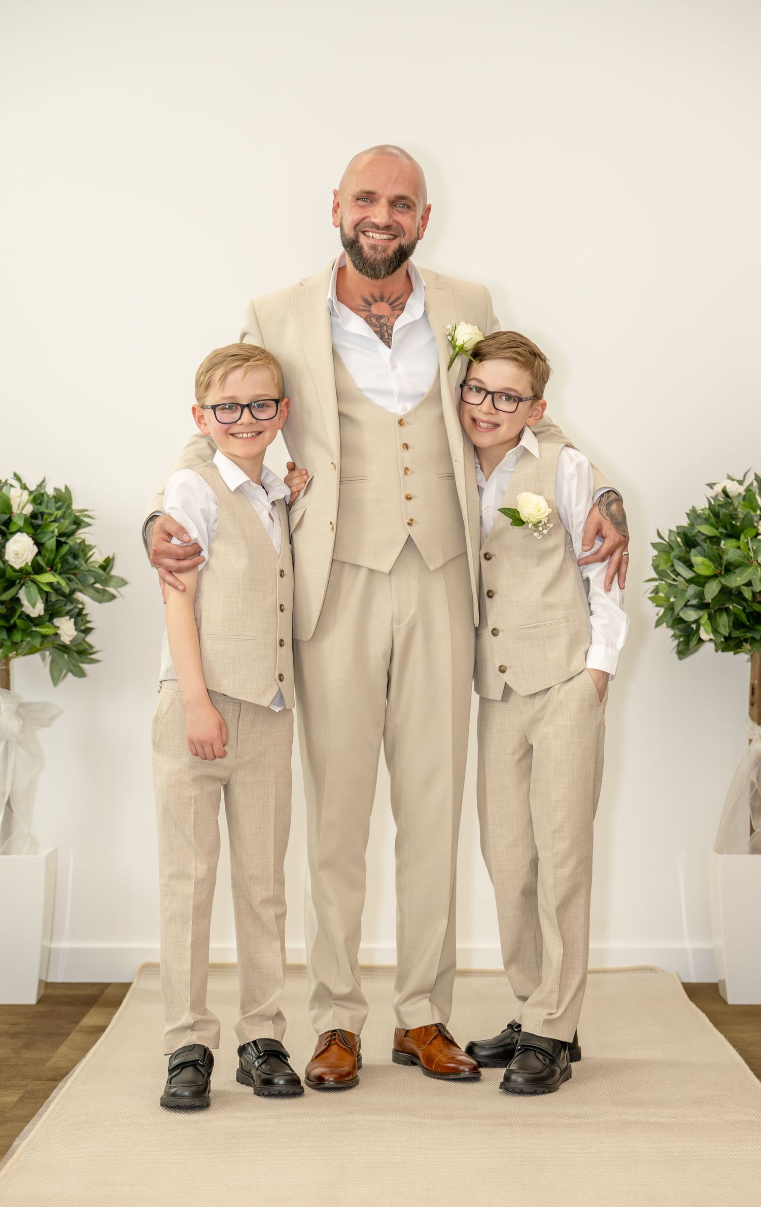 Groom in beige suit posing with two young boys in matching vests at a wedding ceremony.