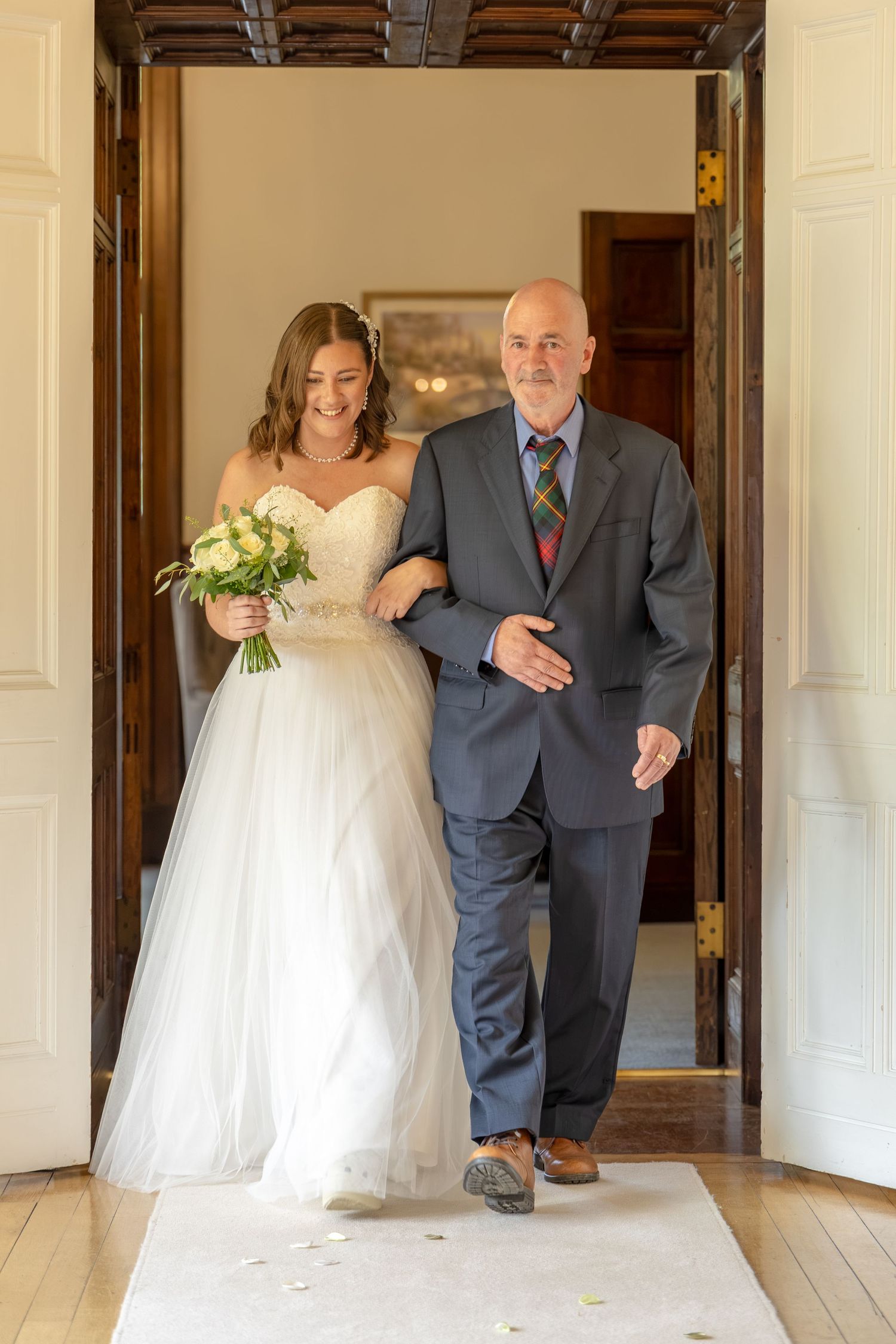 Bride in white gown walks down the aisle with older man in gray suit during elegant wedding ceremony.