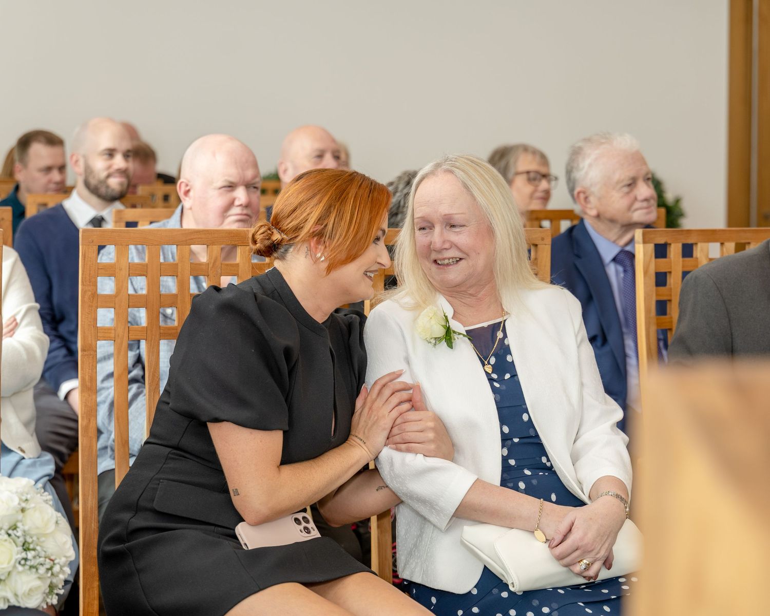 Two women share a warm emotional moment while seated together among guests at a wedding ceremony.