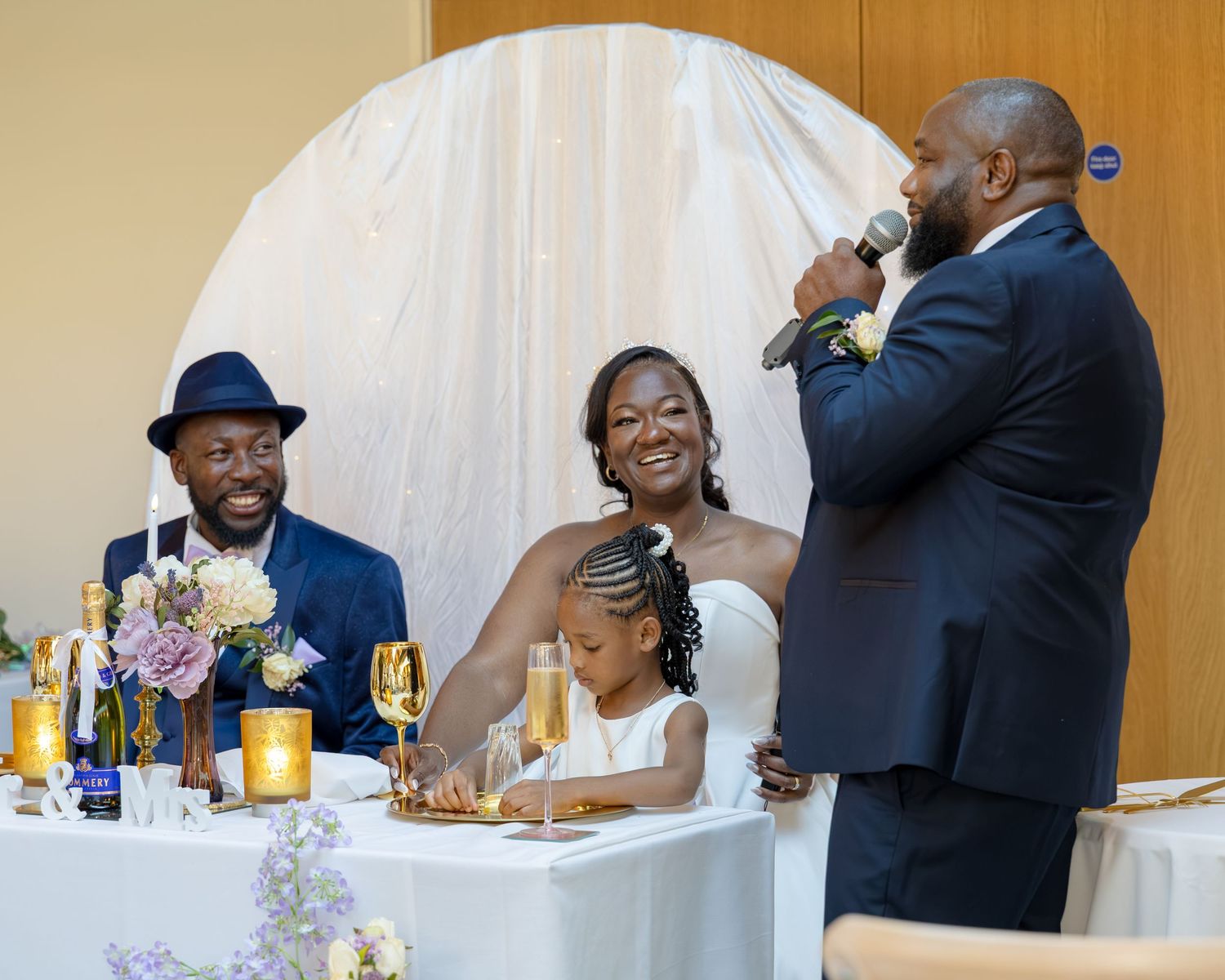 A man gives a wedding toast speech to a smiling bride and groom seated at a decorated sweetheart table.