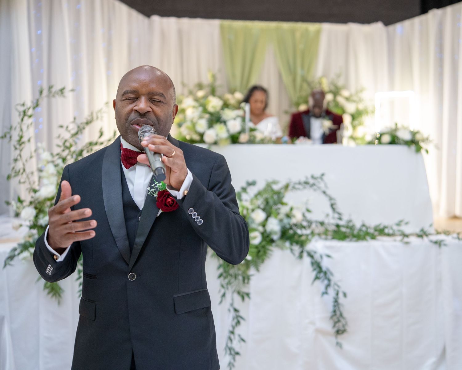Man in tuxedo with bow tie speaks at microphone during elegant wedding reception with floral decorations.