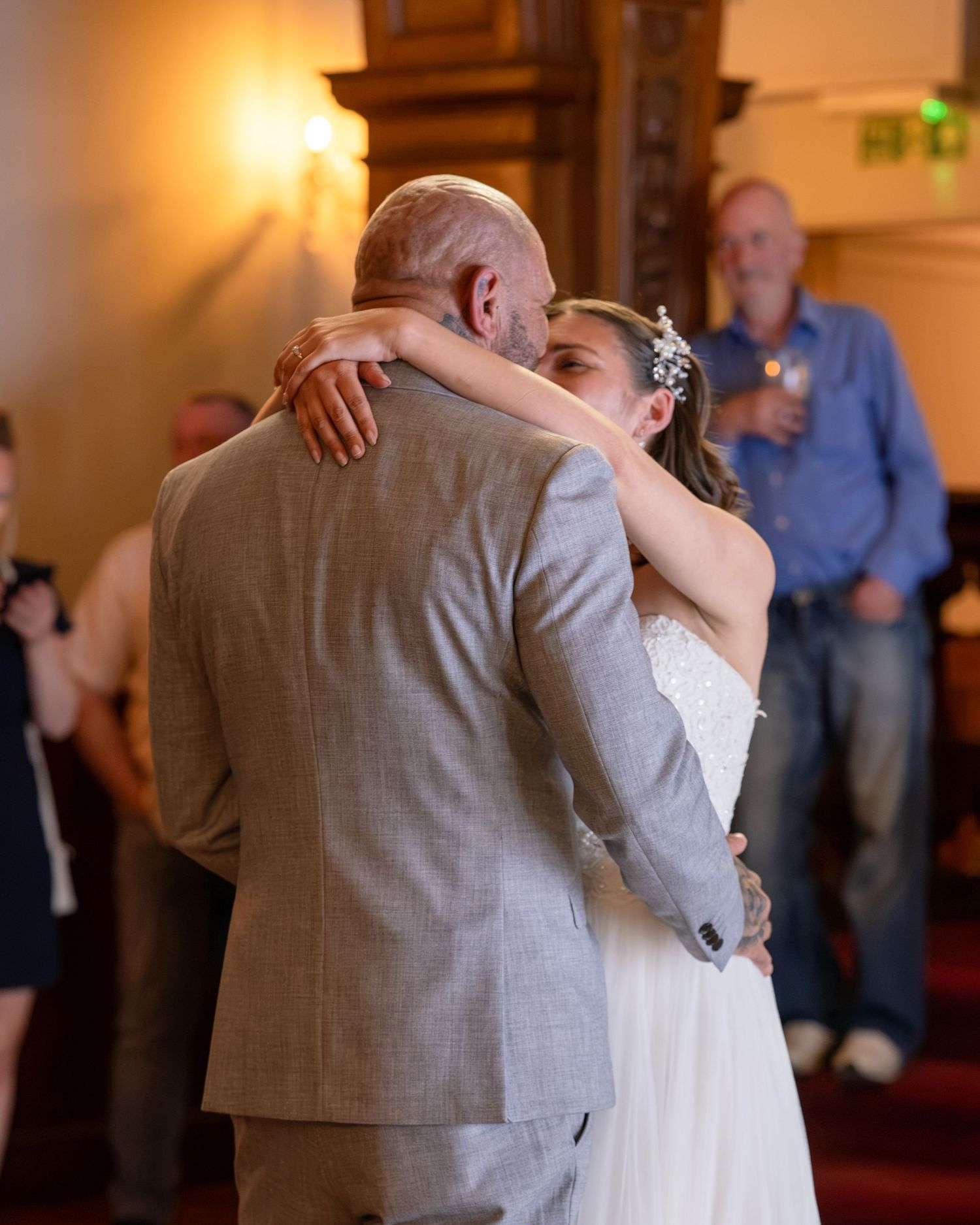 Bride and groom share their first dance embrace at a wedding reception, guests watching in the background.