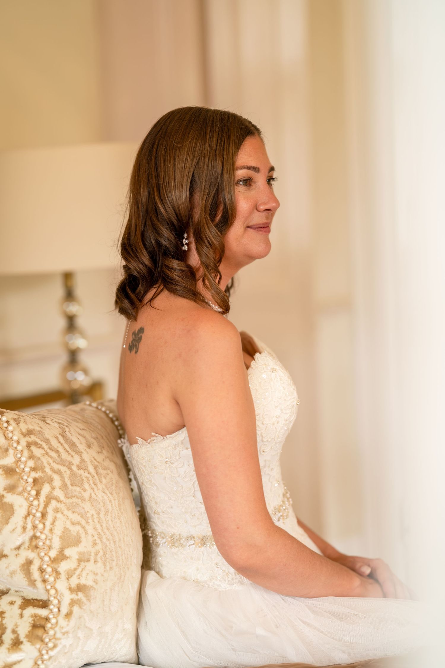 Bride in strapless lace wedding gown with shoulder tattoo sits gracefully on a sofa, smiling softly in a warmly lit room.