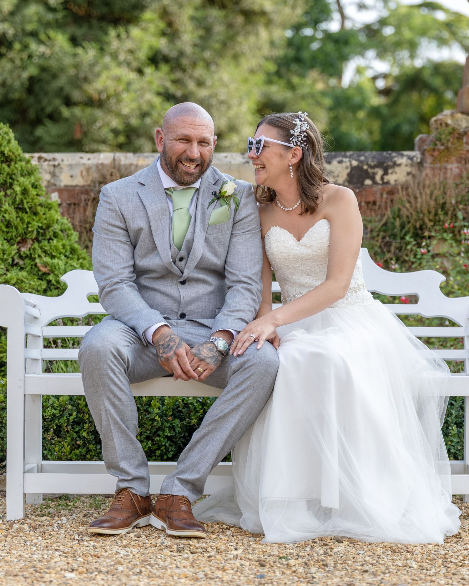 Happy newlyweds sit on a white garden bench, groom in gray suit and bride in white strapless gown with floral hair accessory.