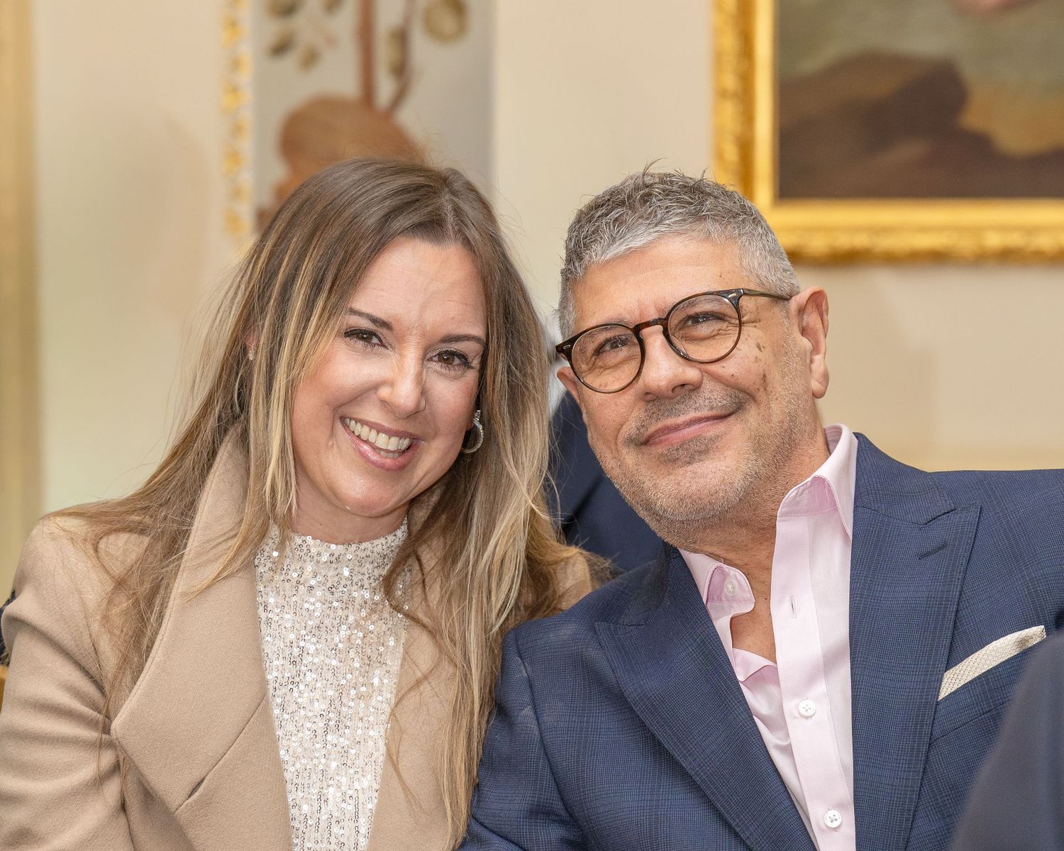 Smiling couple at elegant venue, woman in beige blazer and man with glasses in blue suit posing together.