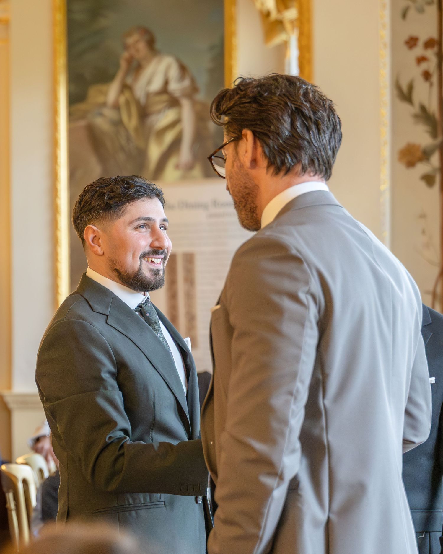 Two men in suits smiling and conversing at a formal wedding ceremony indoors.