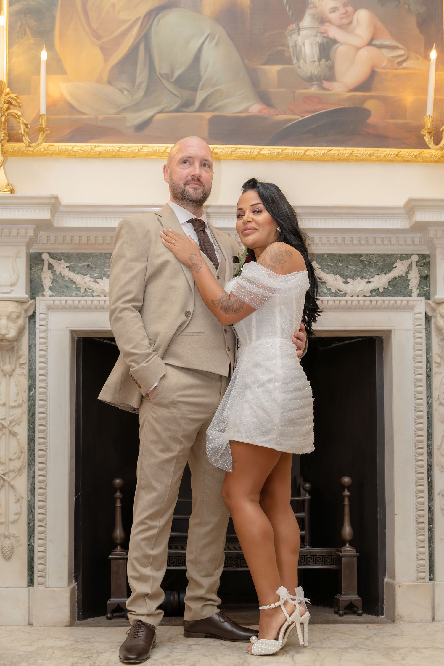 Newlywed couple posing elegantly before grand fireplace with classical painting, bride in white mini dress.