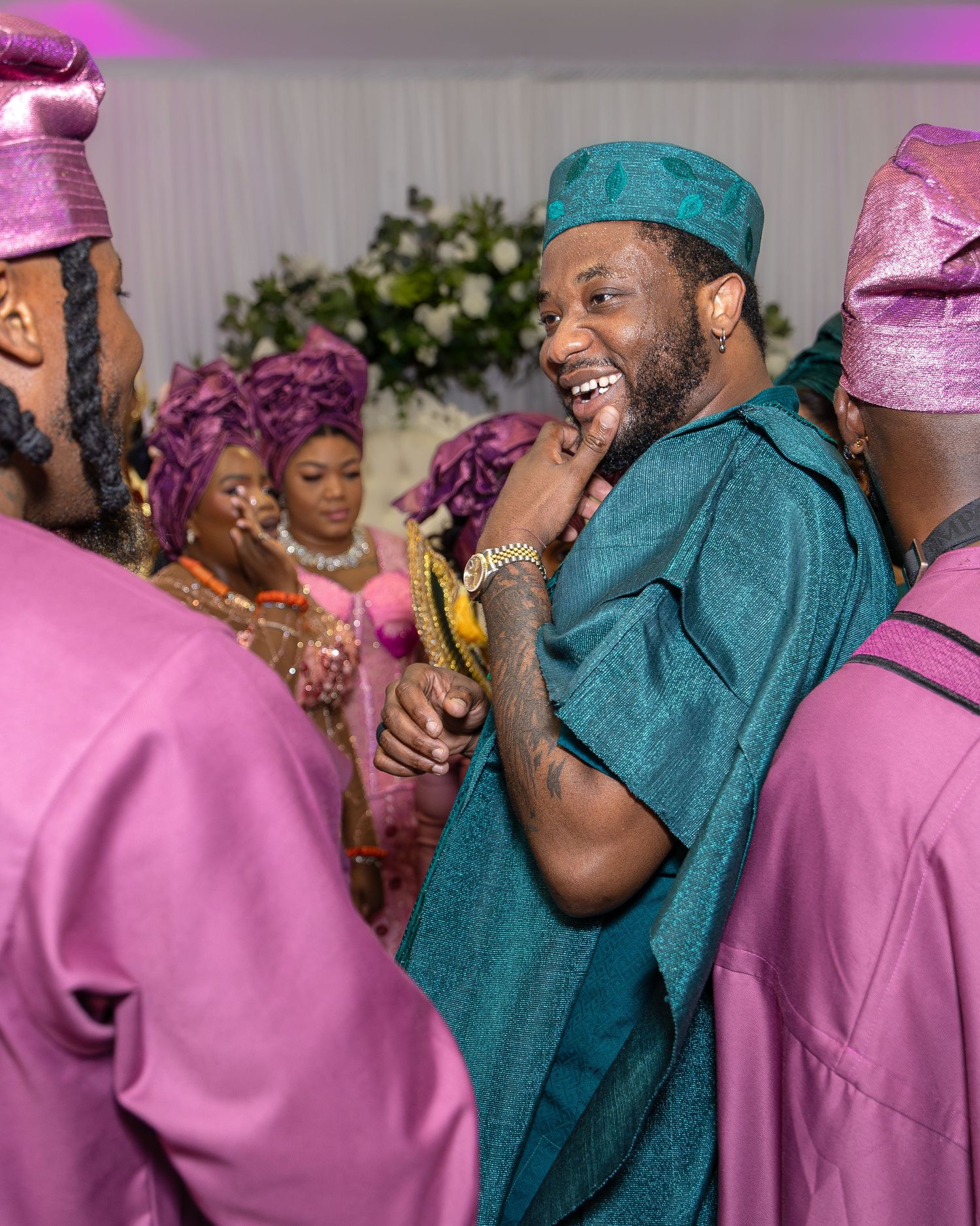 Man in teal agbada laughing with guests in matching pink outfits at vibrant Nigerian traditional wedding reception.