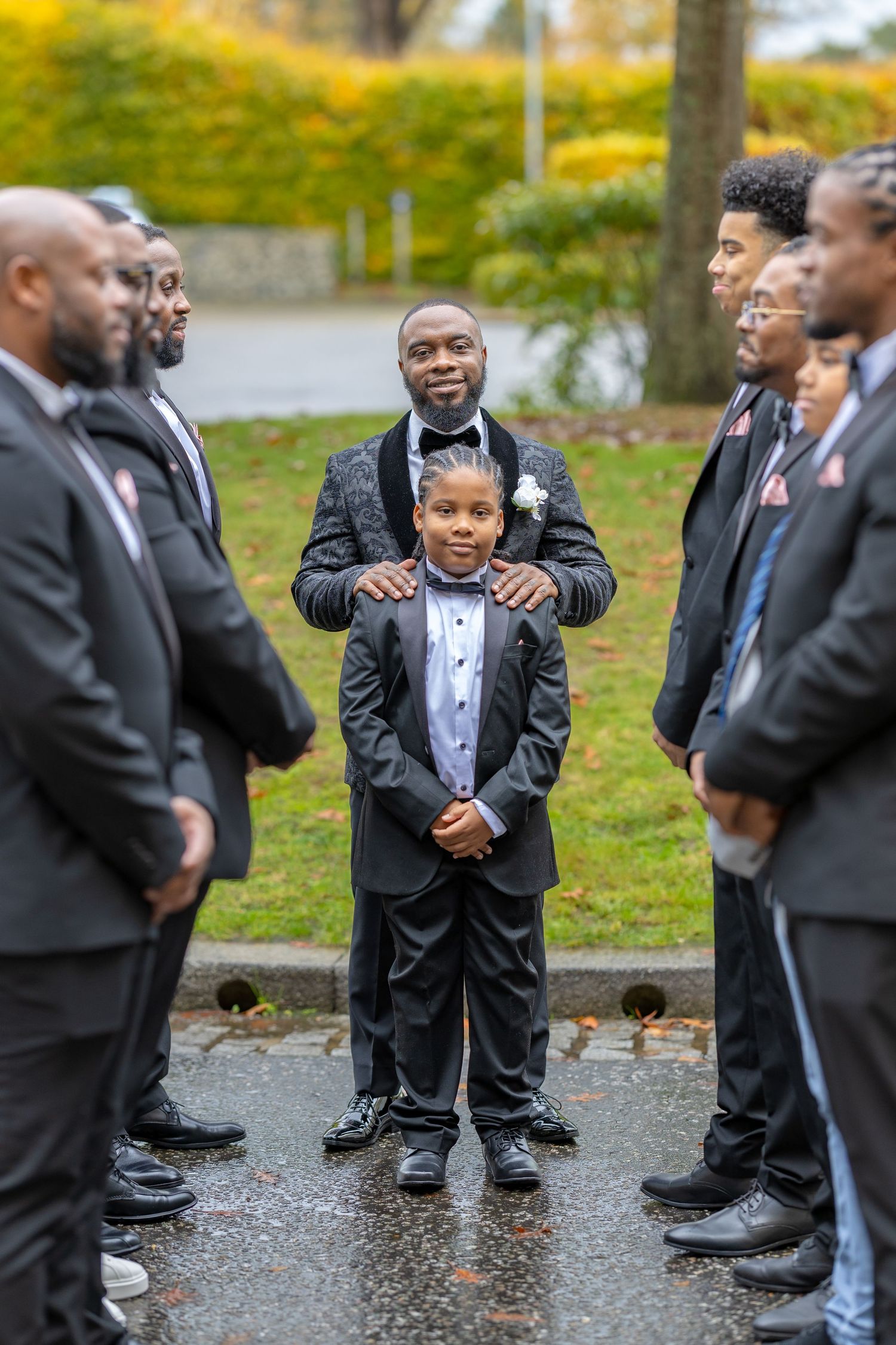 Groom in black tuxedo stands proudly with young ring bearer surrounded by groomsmen outdoors.