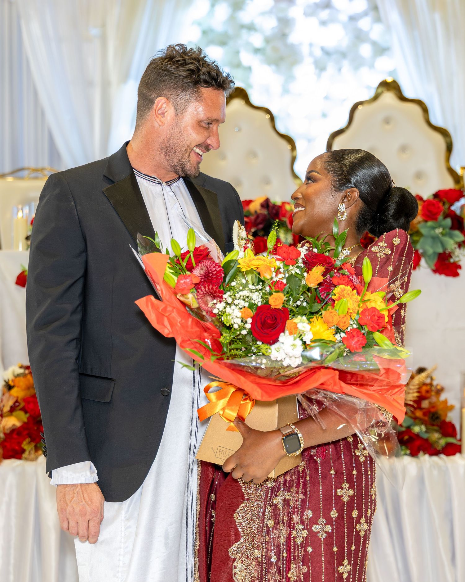 Interracial couple smiling and holding colorful flower bouquet at a traditional African wedding reception.
