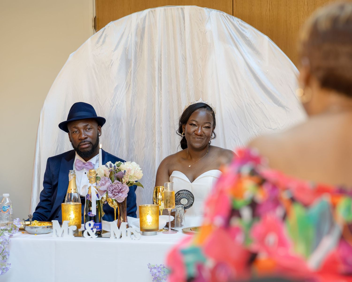 Bride and groom seated at wedding reception sweetheart table with gold decor and Mr & Mrs sign.