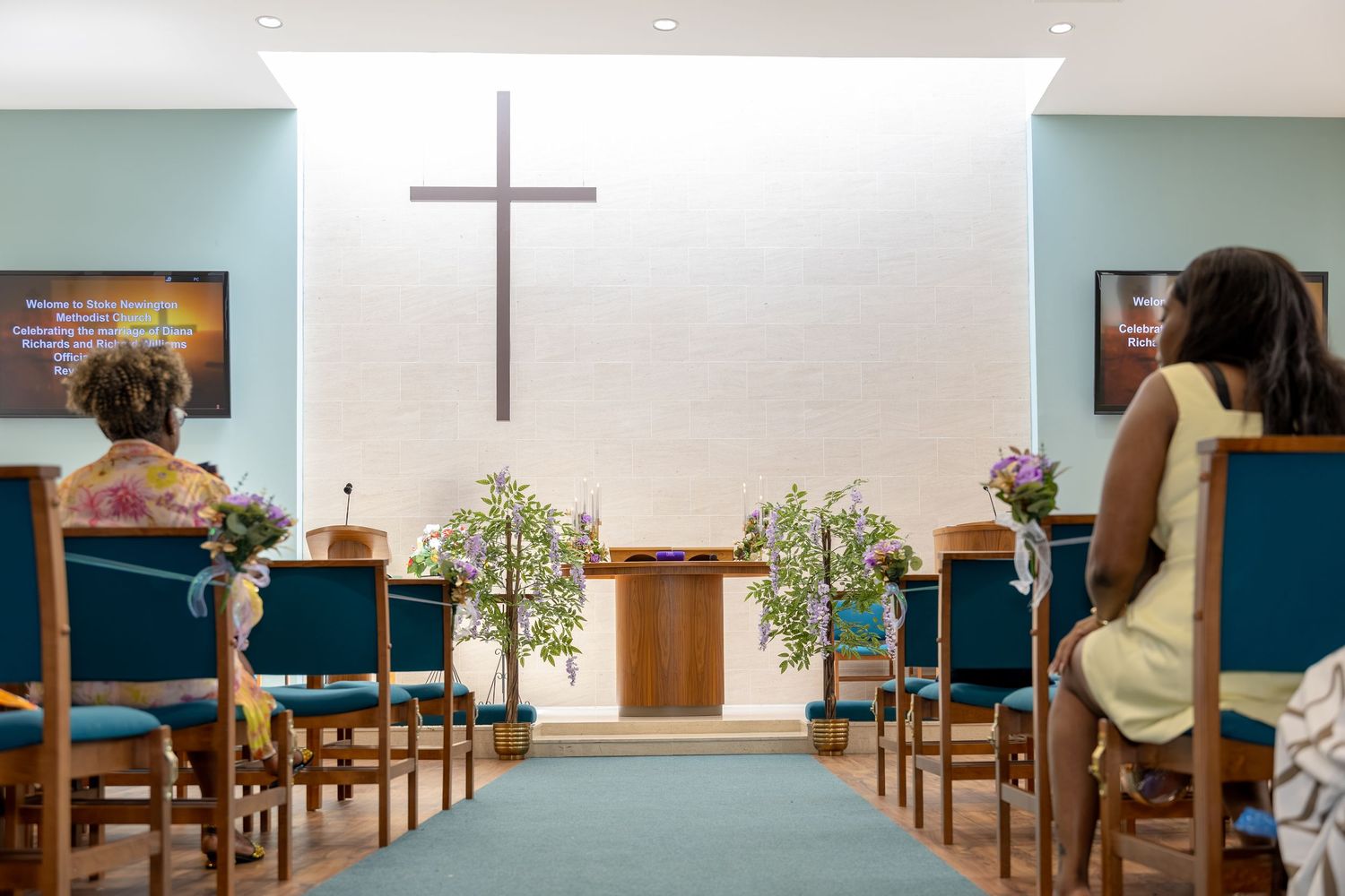 Small church interior with wooden cross, teal pews, floral arrangements, and congregation members seated during a service.