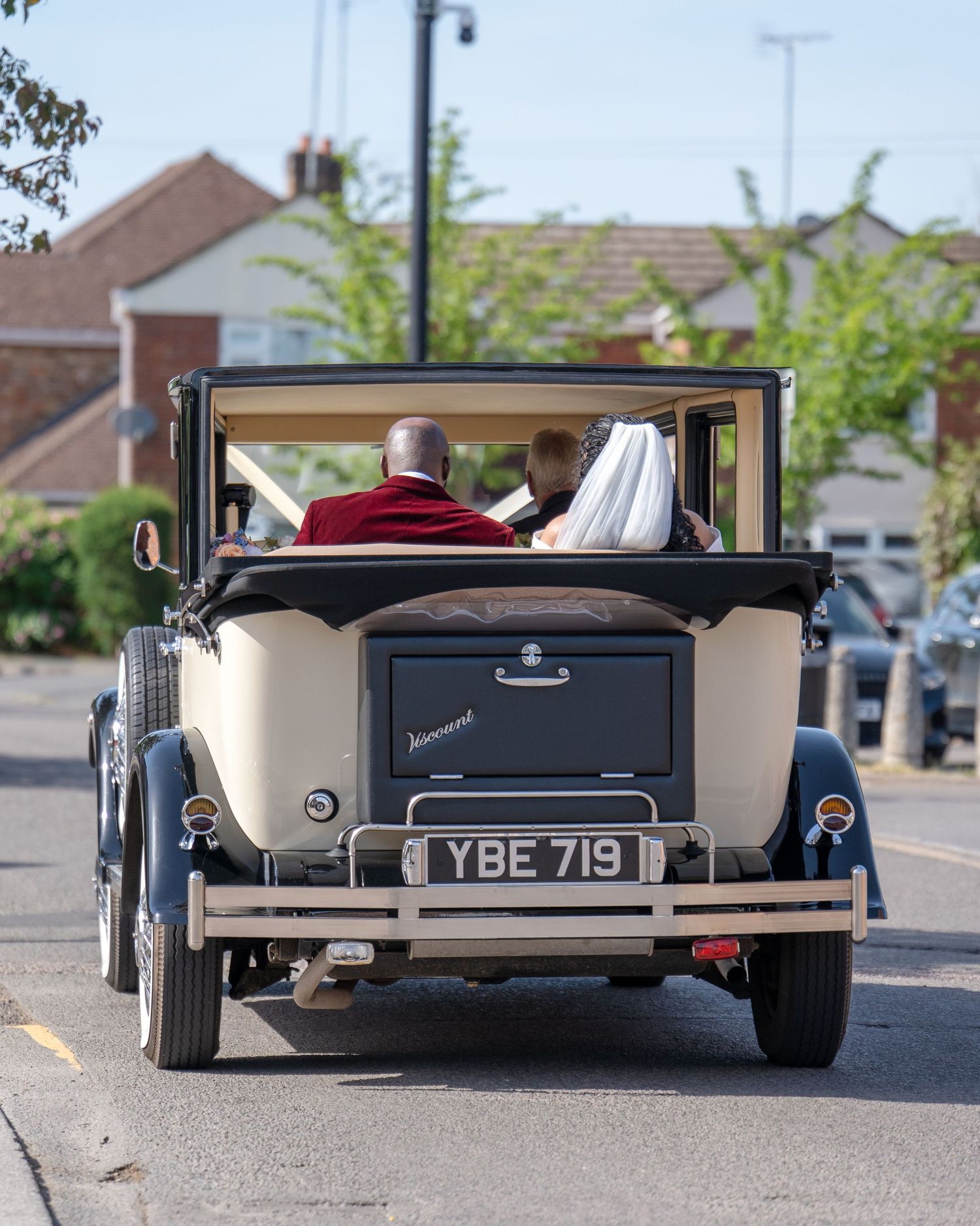 Vintage wedding car with bride and groom driving away, classic convertible with license plate YBE 719 on a sunny day.