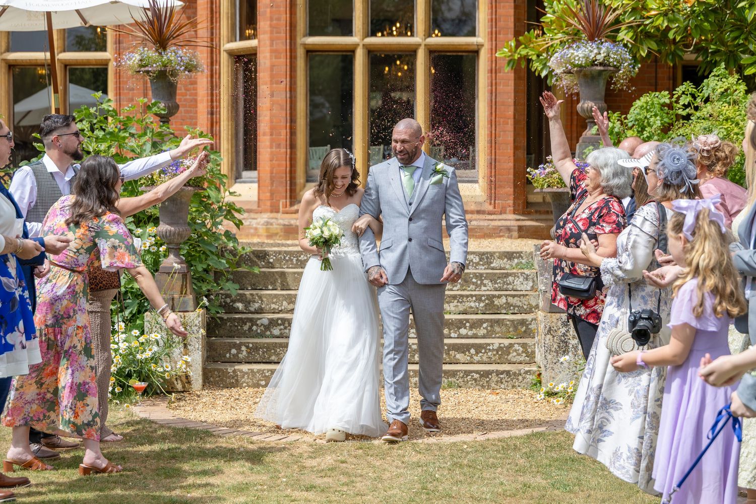 Bride and groom walk through confetti line outside elegant brick venue on sunny wedding day.