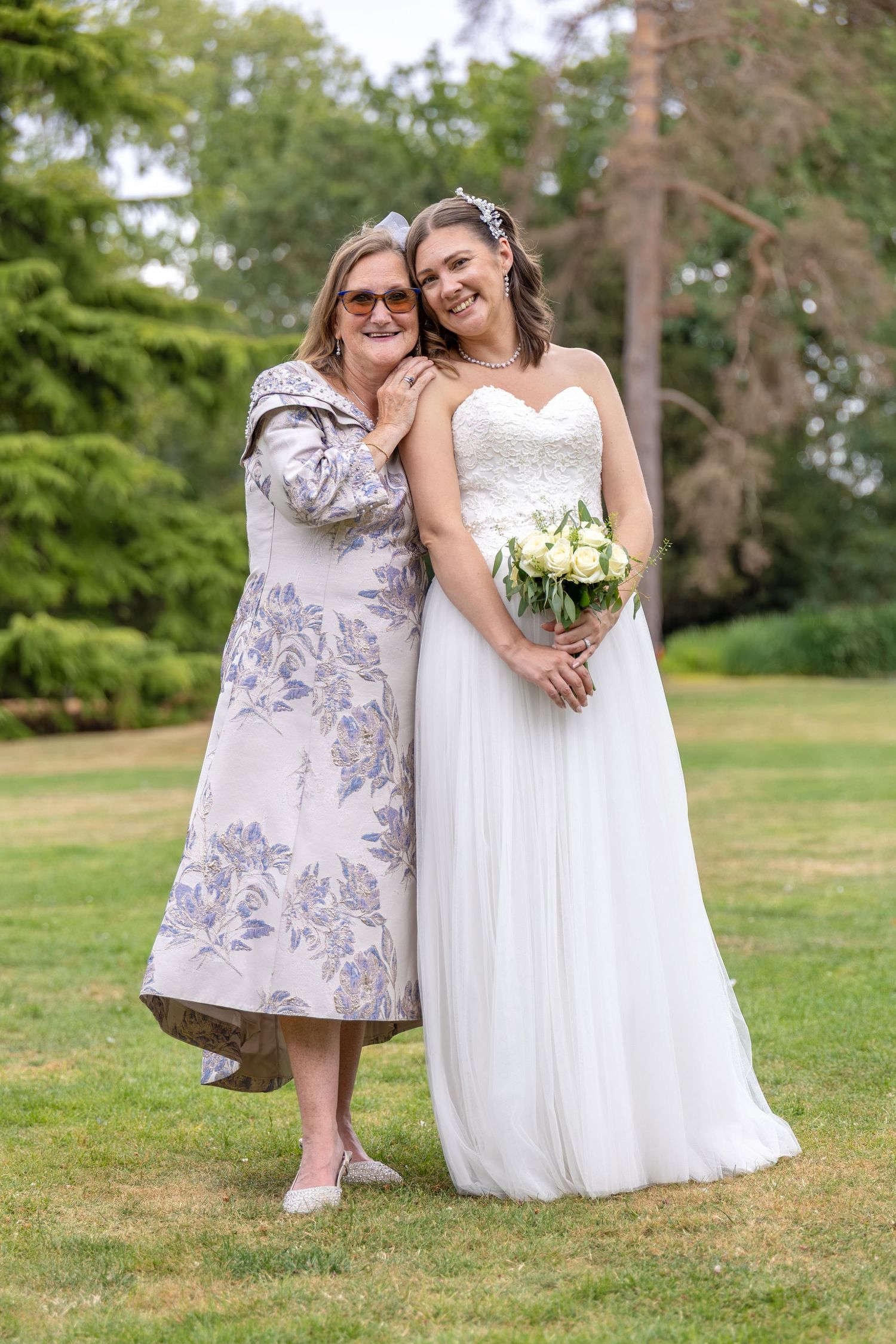 Bride in white gown holding bouquet poses with older woman in floral dress outdoors on green lawn.