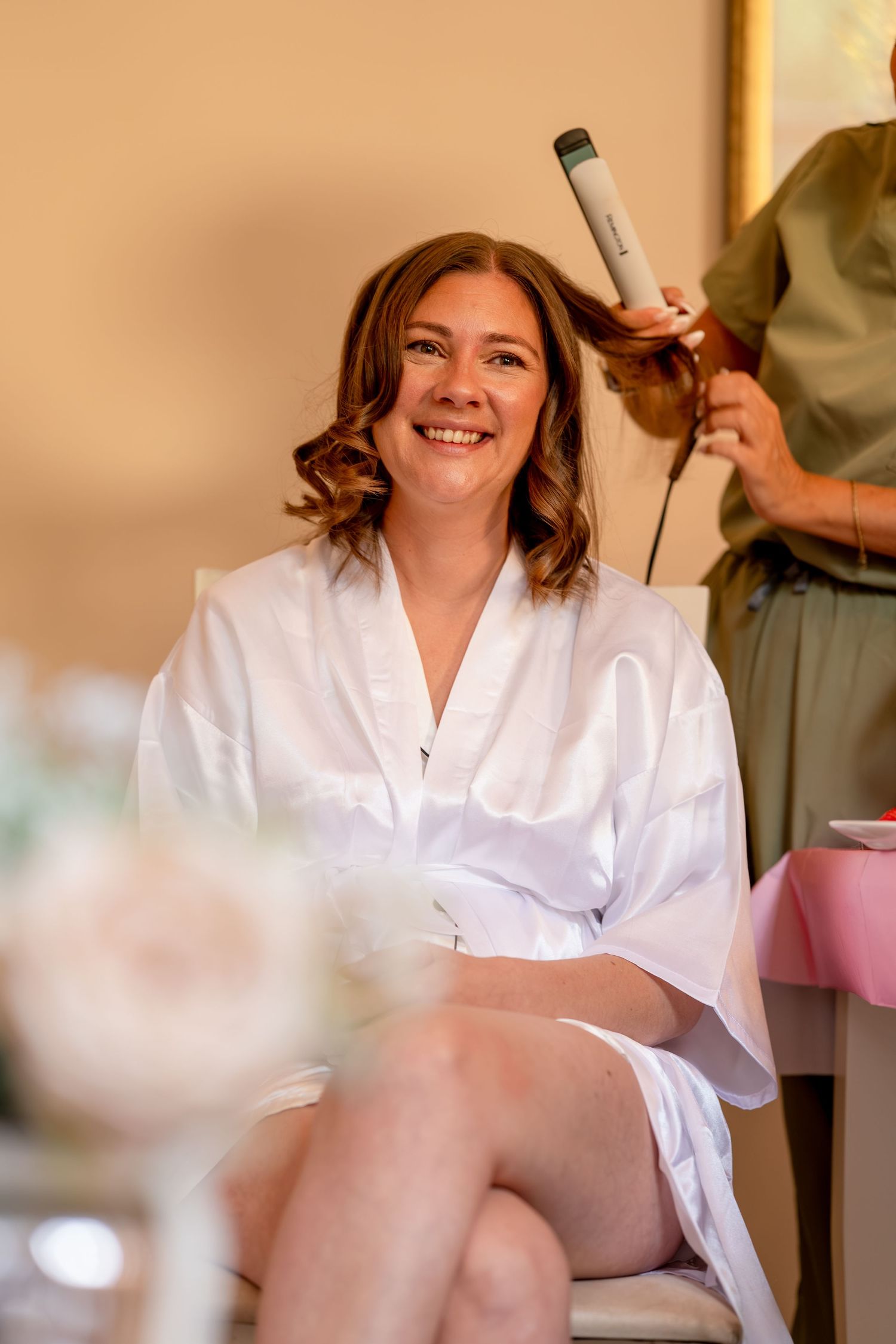 Smiling bride in white robe getting hair styled with curling iron during wedding preparation.