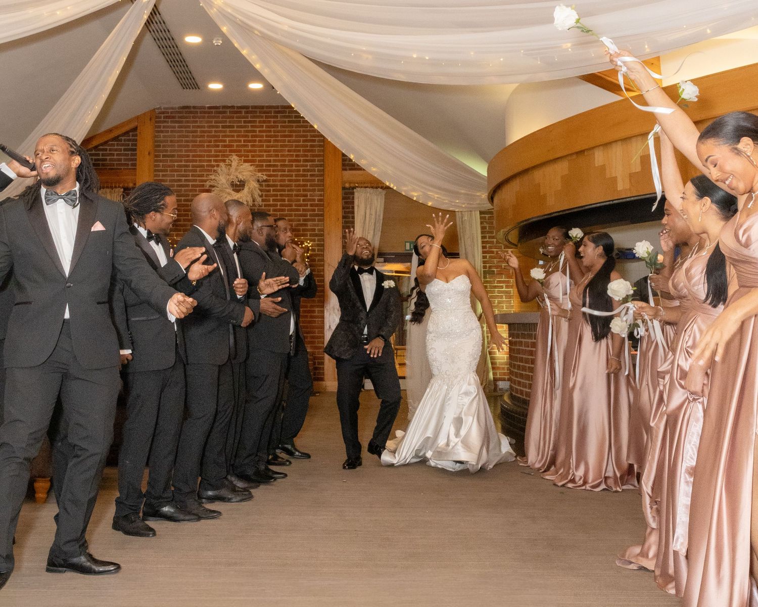 Bride and groom walk through cheering wedding party in elegant venue with draped ceilings and bridesmaids in champagne gowns.