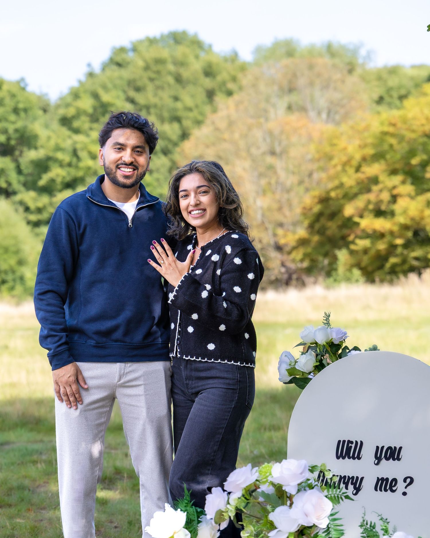 Couple posing happily next to a Will You Marry Me sign after a successful marriage proposal outdoors.