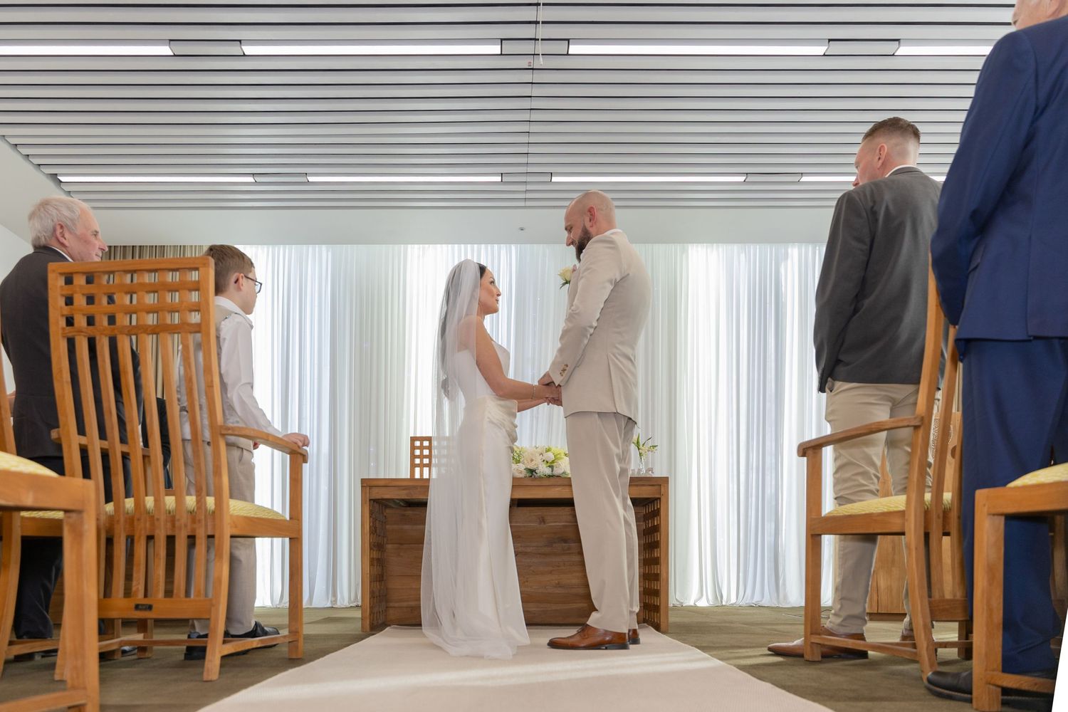 Bride and groom exchange vows at indoor wedding ceremony with guests seated in wooden chairs.