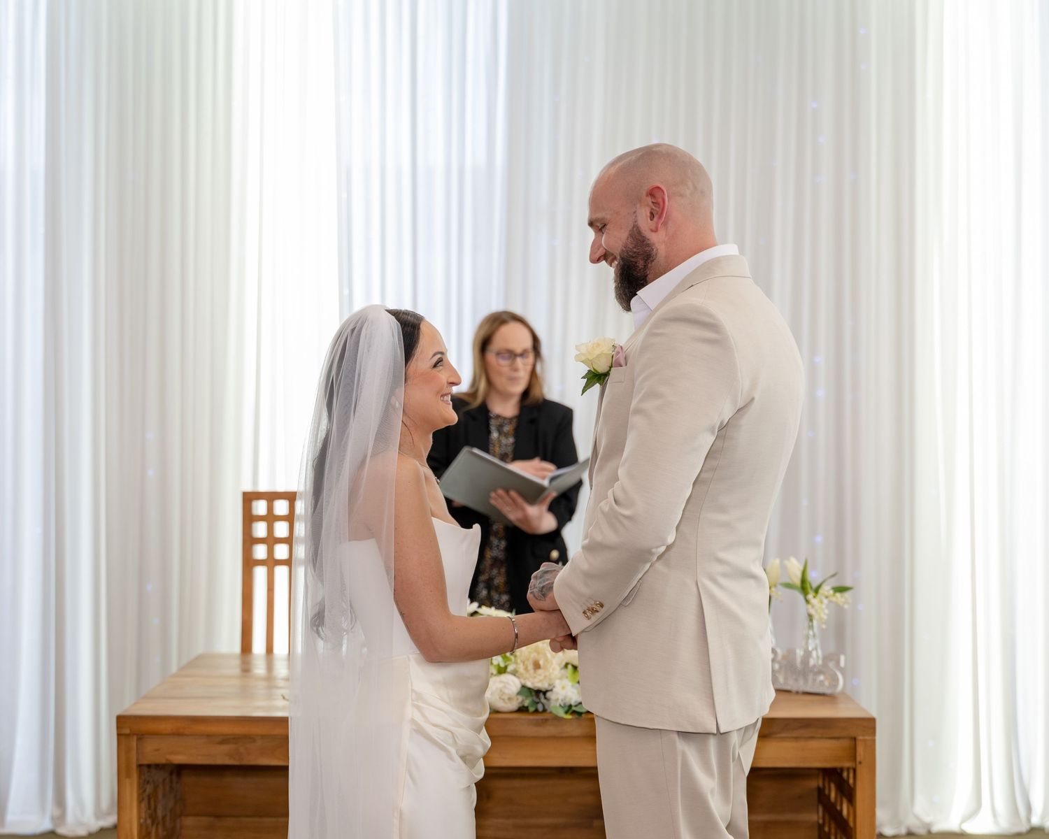 Smiling bride in veil holds hands with groom as officiant reads from book at altar.