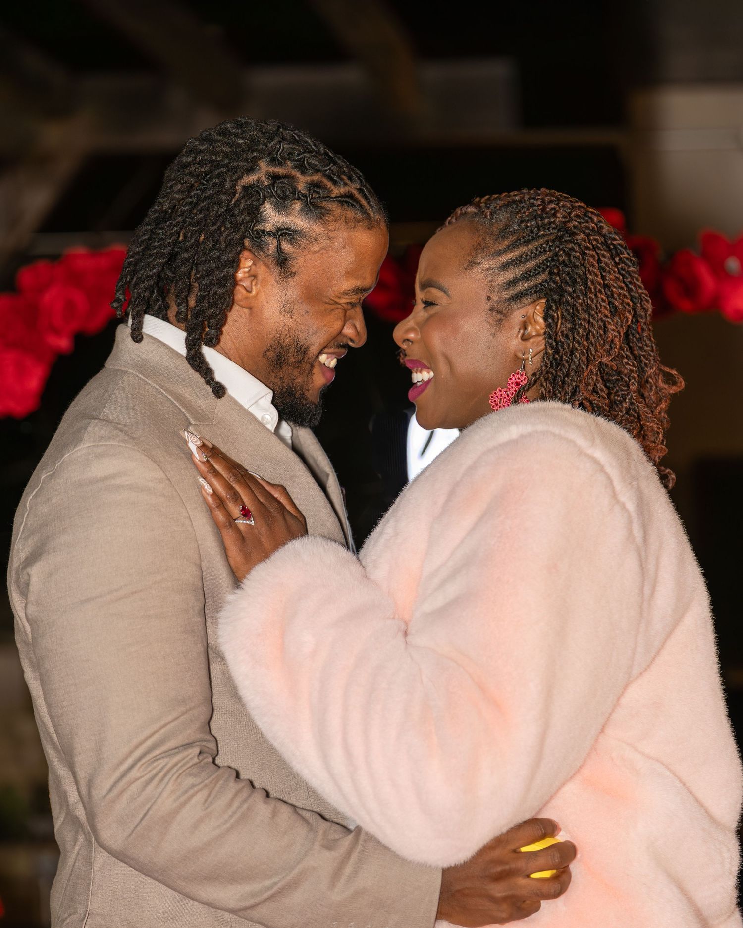 Newly engaged couple smiles joyfully at each other surrounded by red roses after proposal.