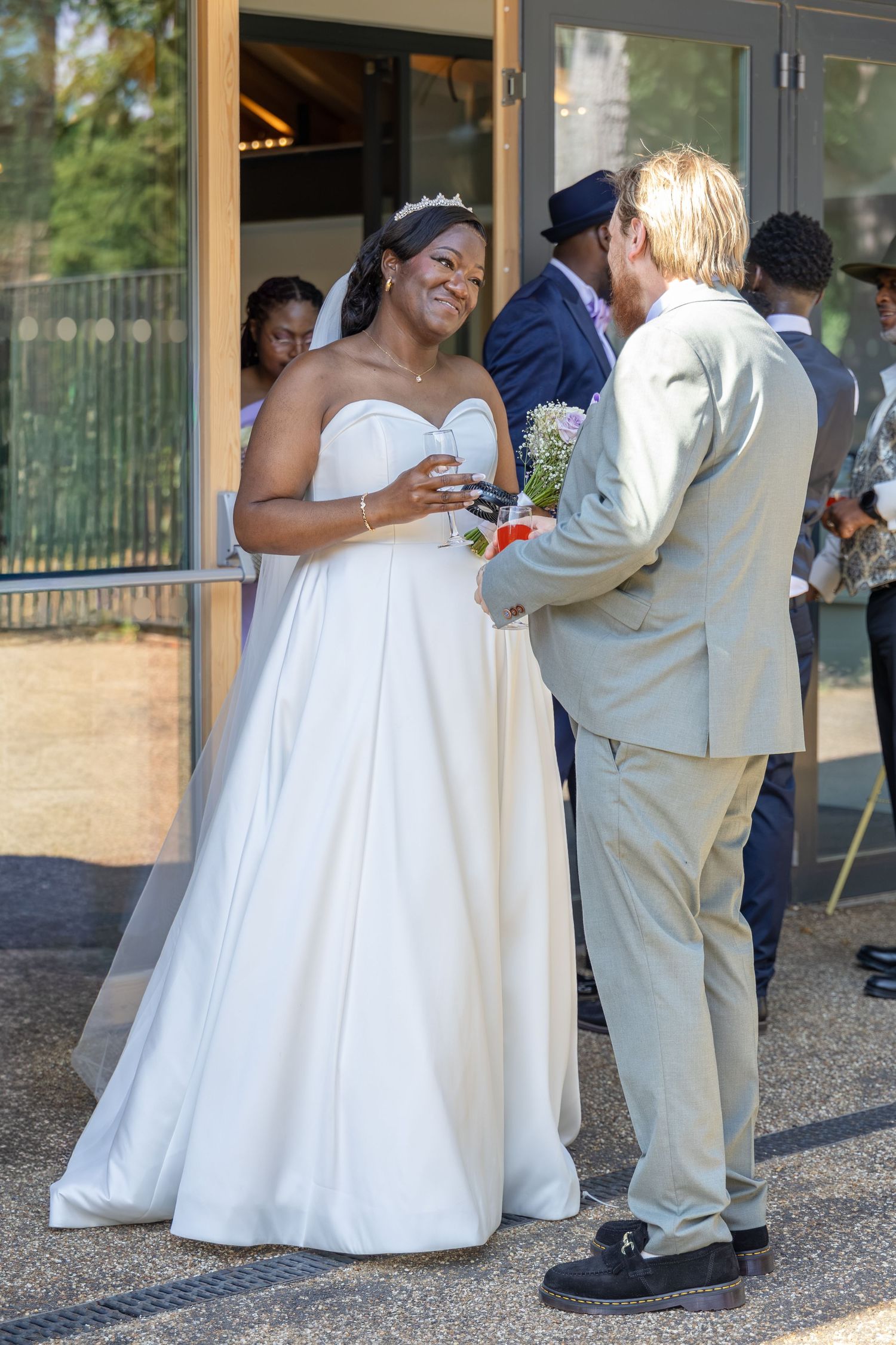 Bride in white gown and groom in tan suit share a moment outside their wedding venue on a sunny day.
