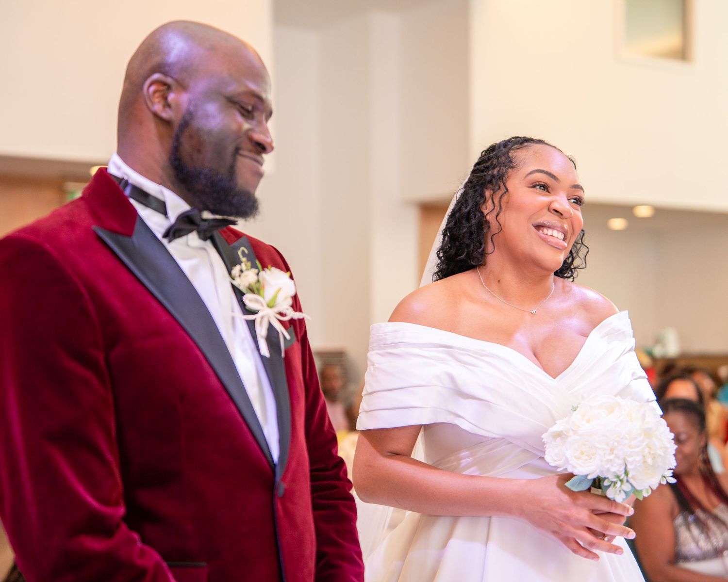 Bride in white off-shoulder gown and groom in burgundy velvet tuxedo smiling joyfully at wedding ceremony.