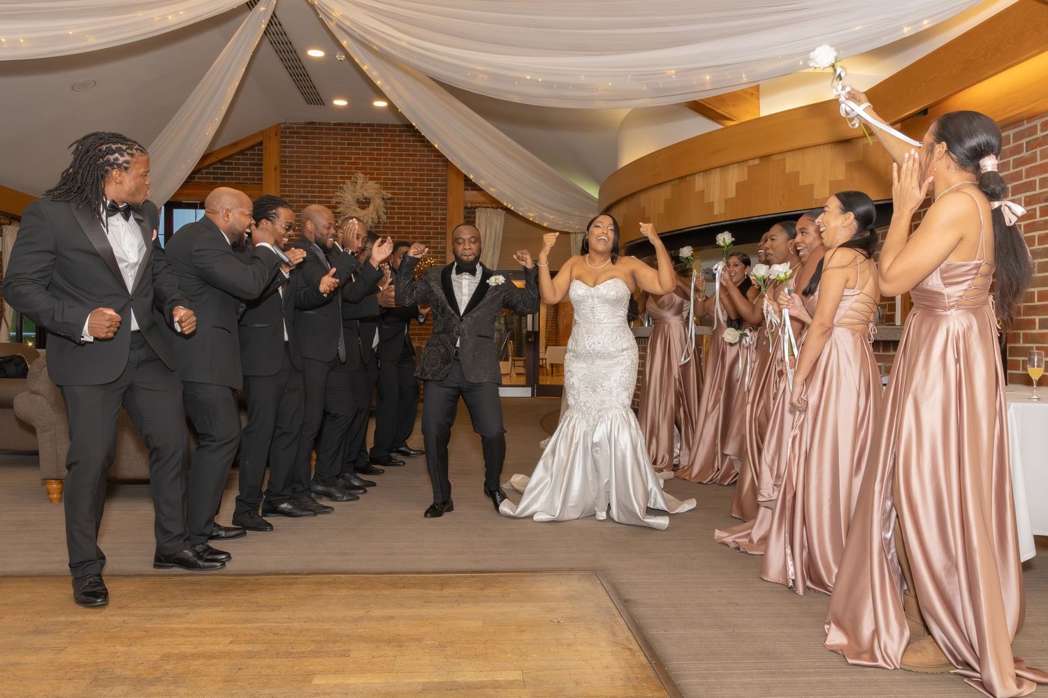 Bride and groom enter wedding reception as bridesmaids in champagne gowns and groomsmen celebrate.