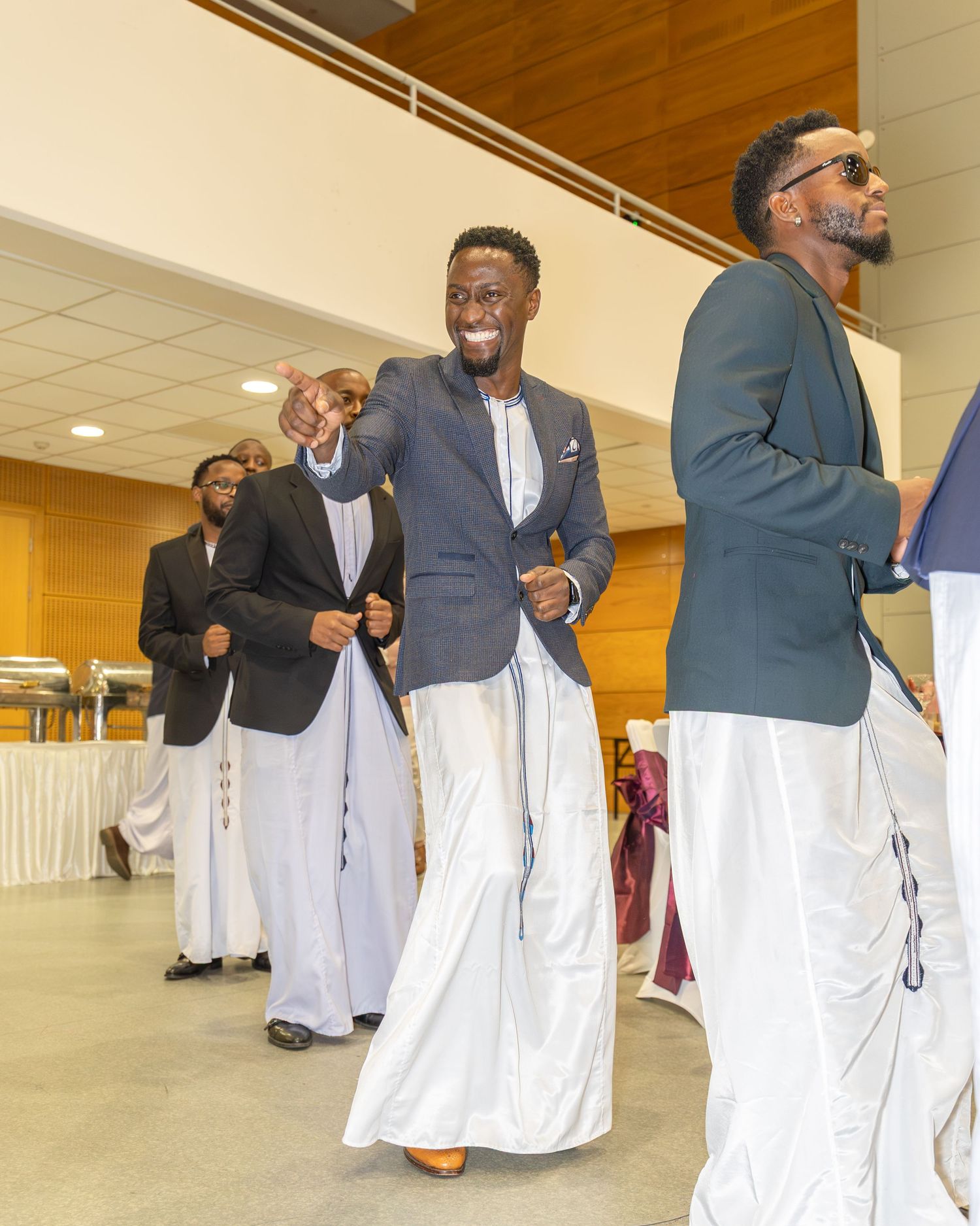 Men in traditional West African white boubou robes and blazers walk and dance at a festive cultural celebration.