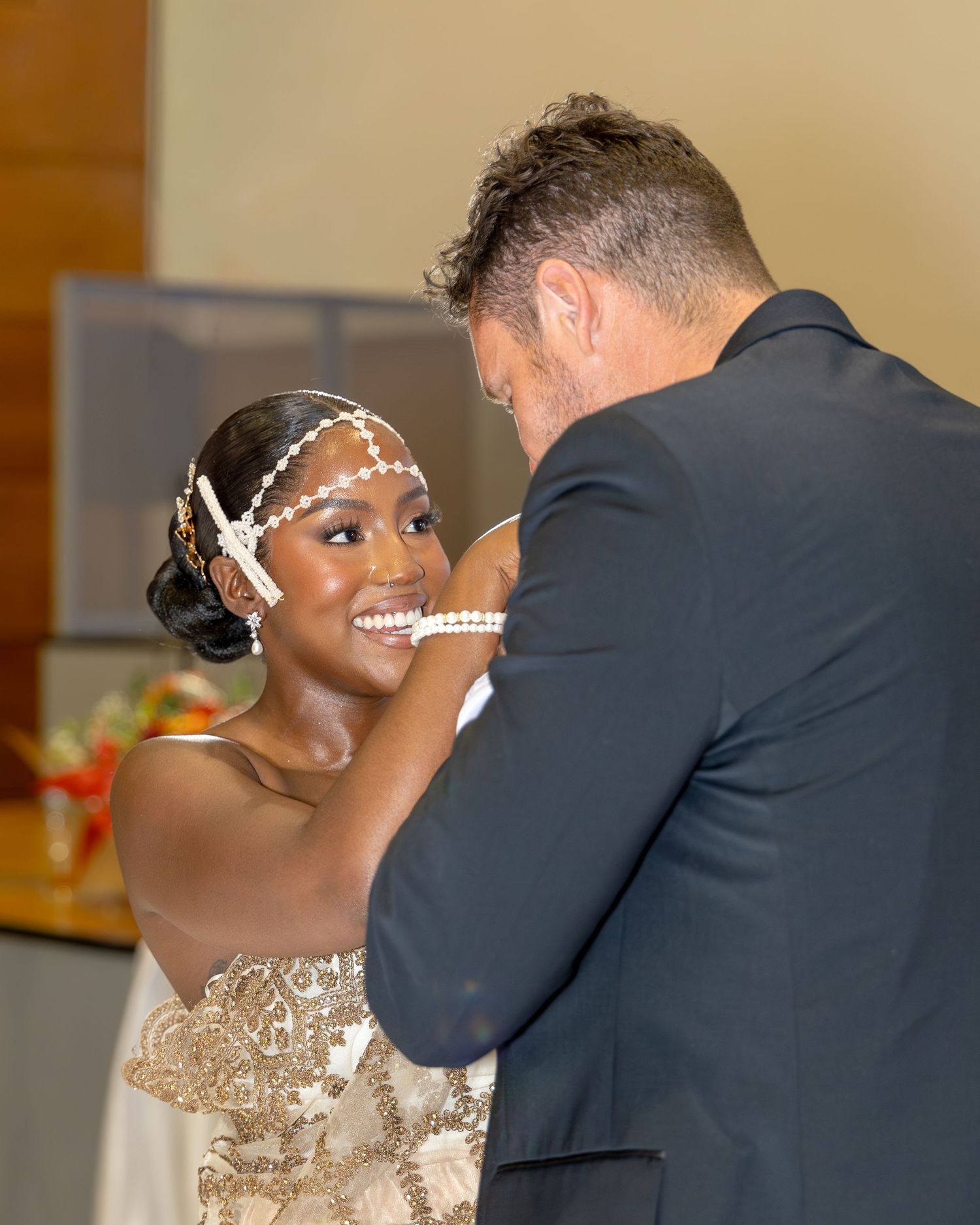 Groom in dark suit tenderly touches bride's face as she smiles in gold beaded gown with pearl headpiece.