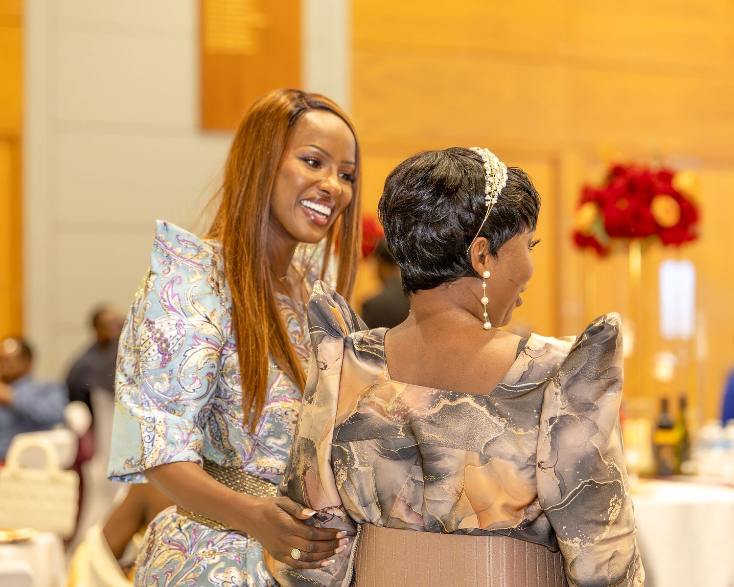 Two elegantly dressed women smiling and embracing at a wedding reception.