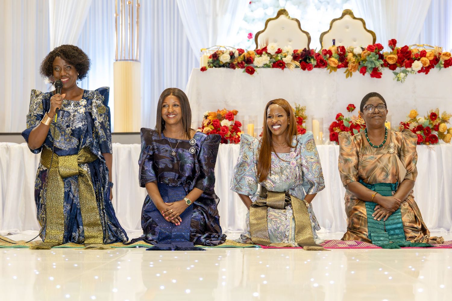 Four women in colorful African traditional attire kneel on stage at a cultural ceremony with floral backdrop.