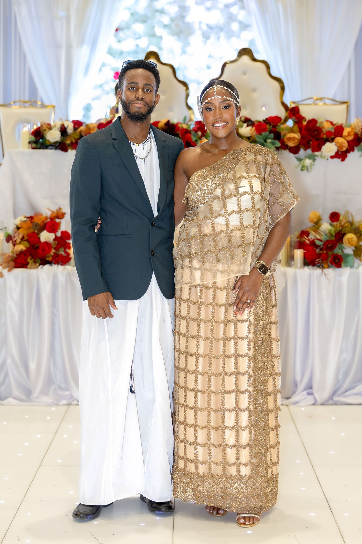 Couple poses together at wedding reception, man in dark blazer and white pants, woman in gold grid dress.