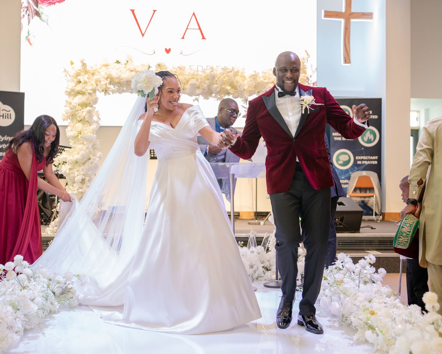 Joyful newlyweds exit church ceremony, bride in white gown, groom in red velvet tuxedo, floral arch backdrop.