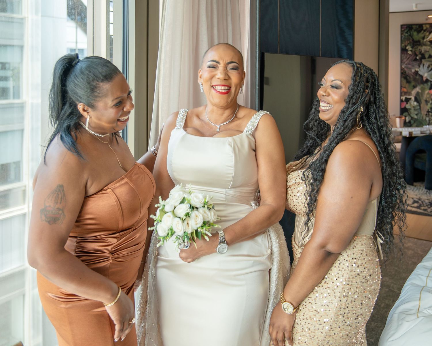 Bride in white gown holding bouquet smiles with two bridesmaids in satin and gold dresses near window.