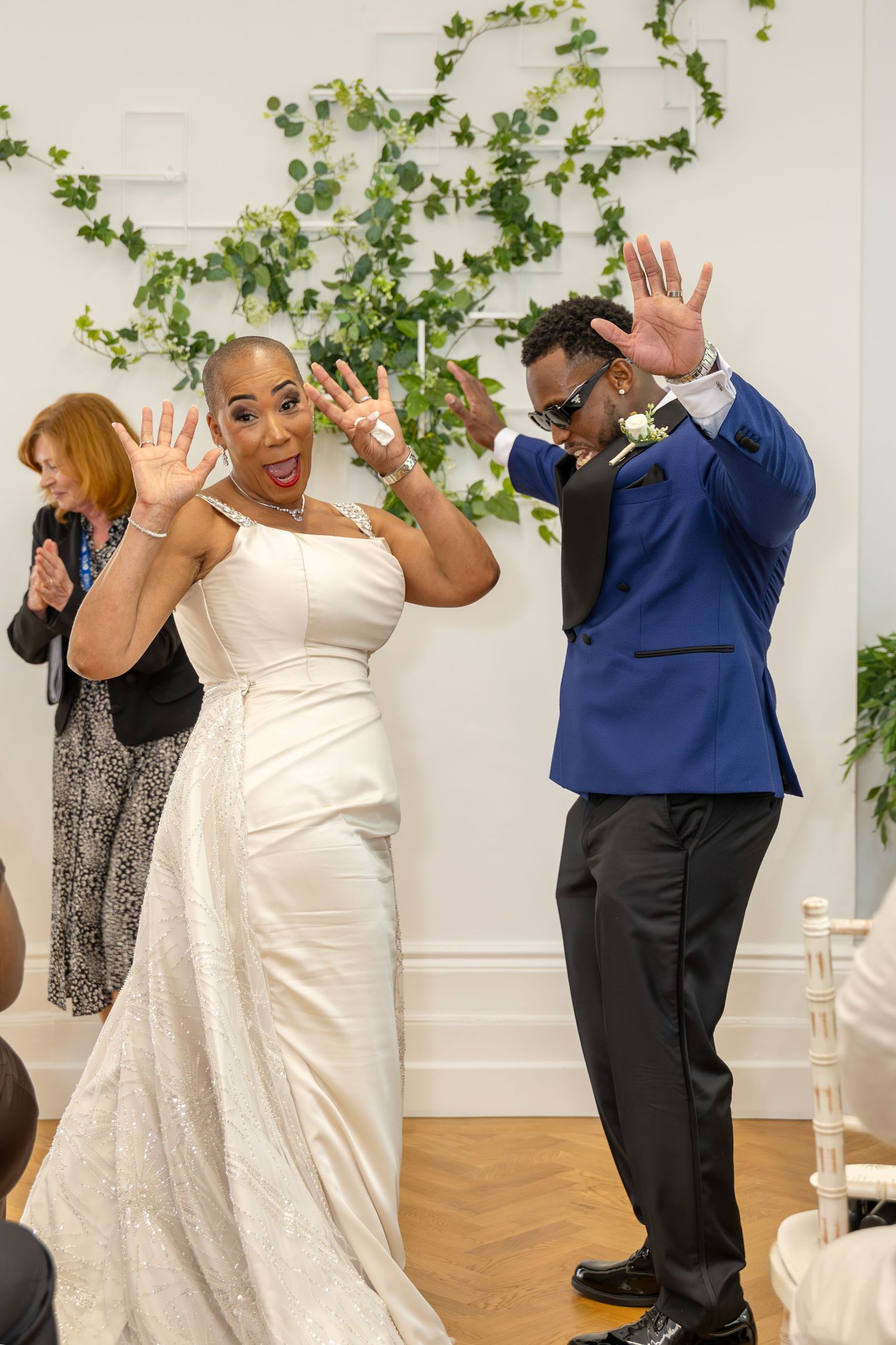 Joyful bride in white gown and groom in blue suit celebrating at wedding reception with ivy wall backdrop.
