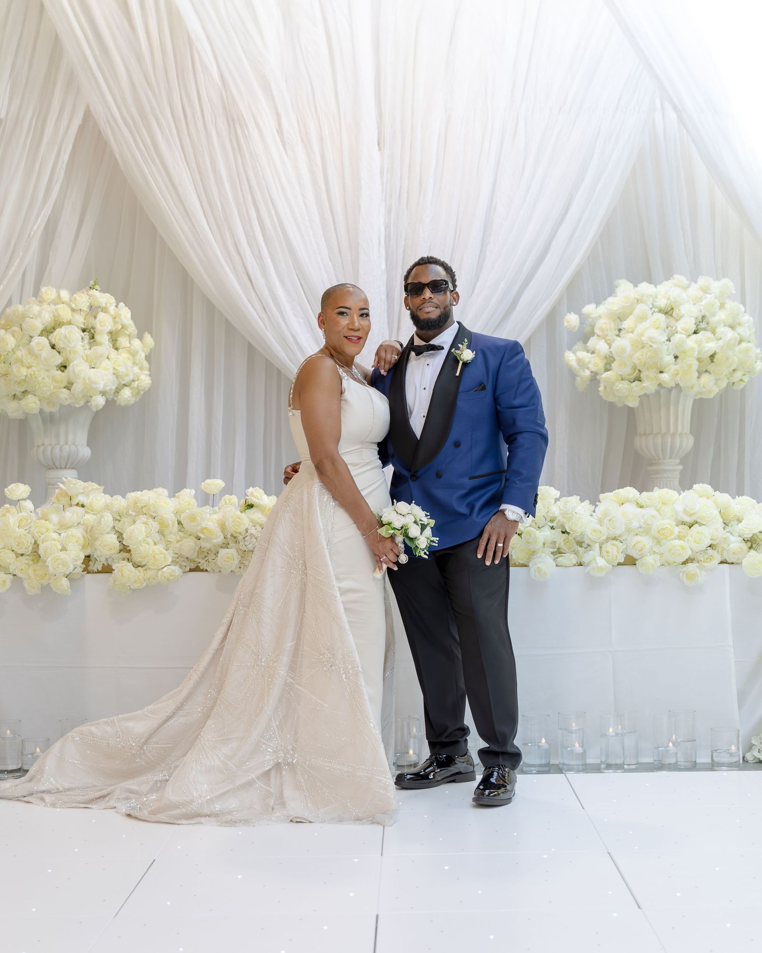 Elegant wedding couple posing before white floral arrangements and draped backdrop, bride in gown, groom in blue suit.