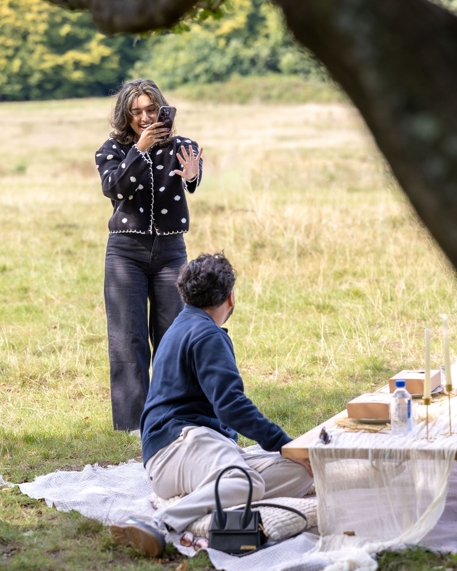 Woman admiring engagement ring while man sits at decorated picnic proposal setup in grassy field.