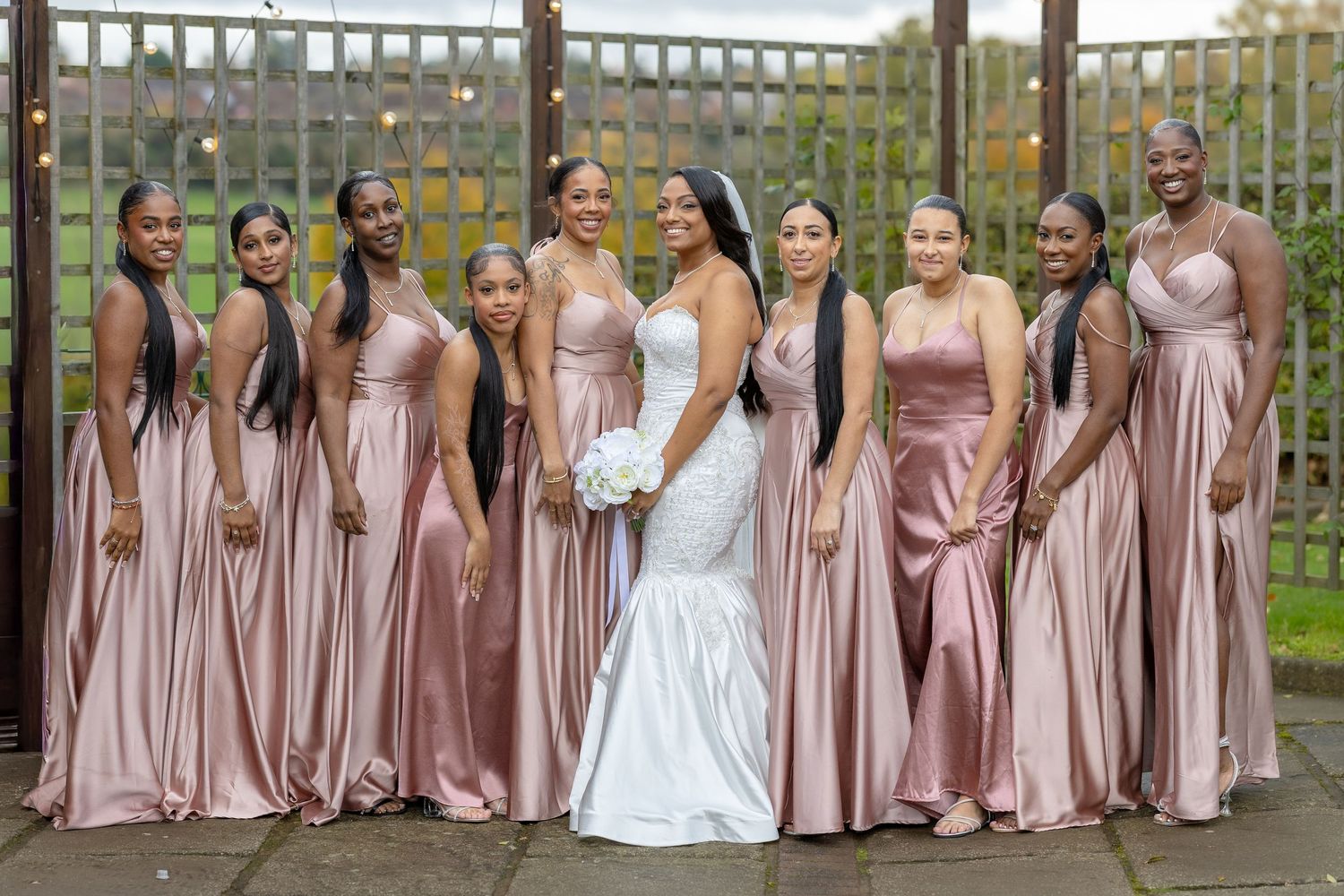 Bride in white mermaid gown stands with nine bridesmaids wearing matching dusty rose satin dresses at outdoor wedding.
