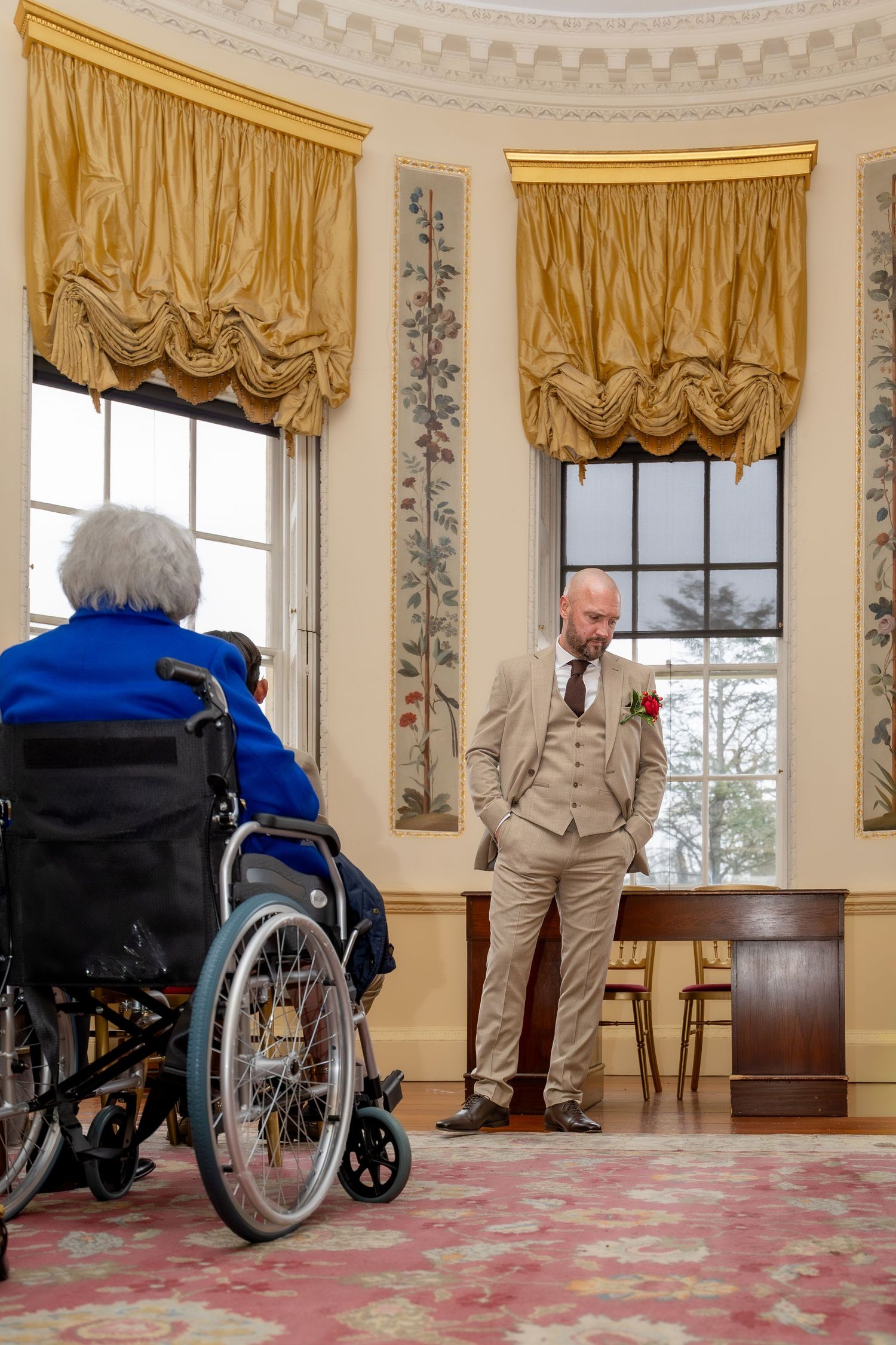 Bald groom in beige three-piece suit with red boutonniere standing near elderly woman in wheelchair.