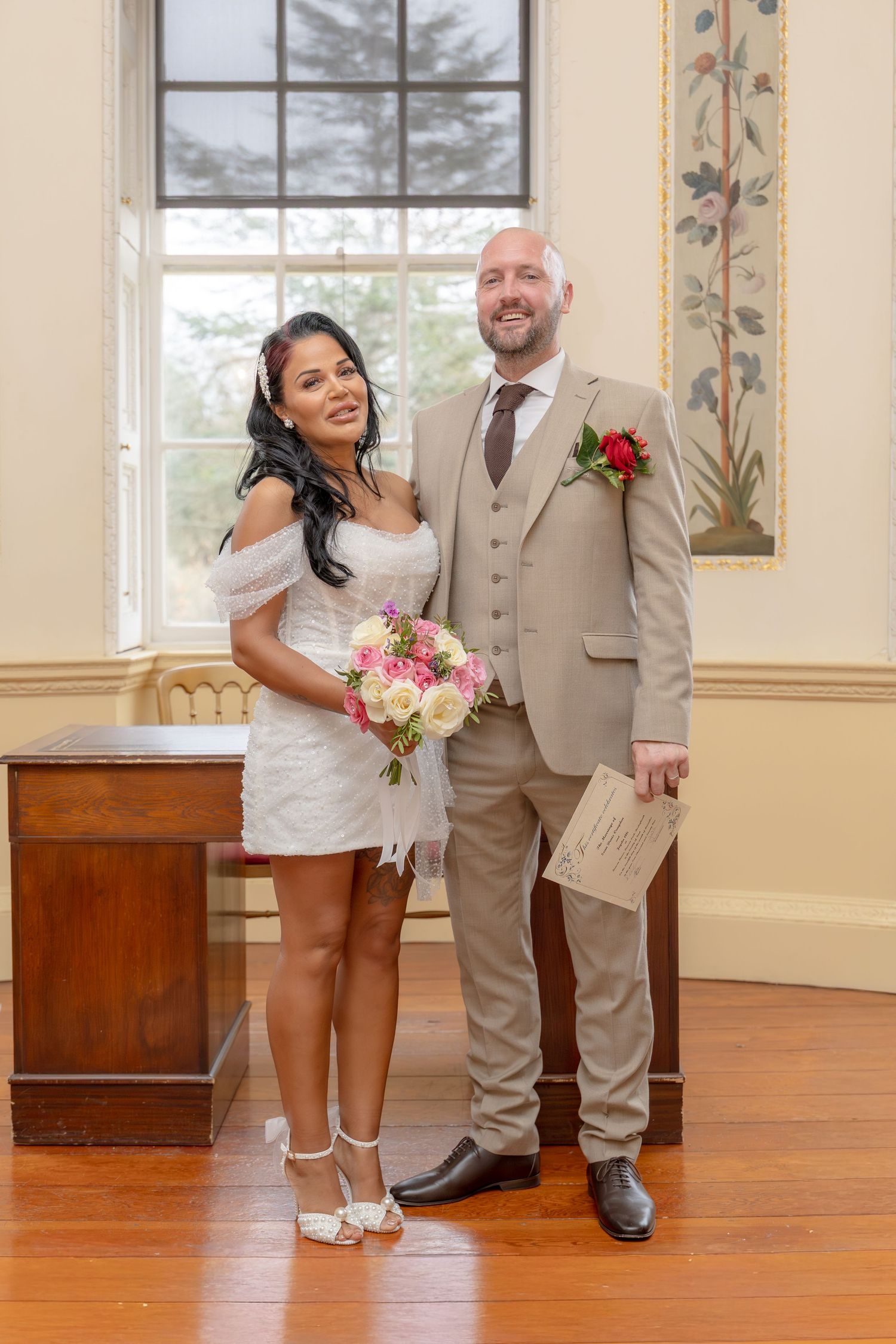 Smiling couple pose in elegant room, bride with bouquet in white dress, groom in beige suit.