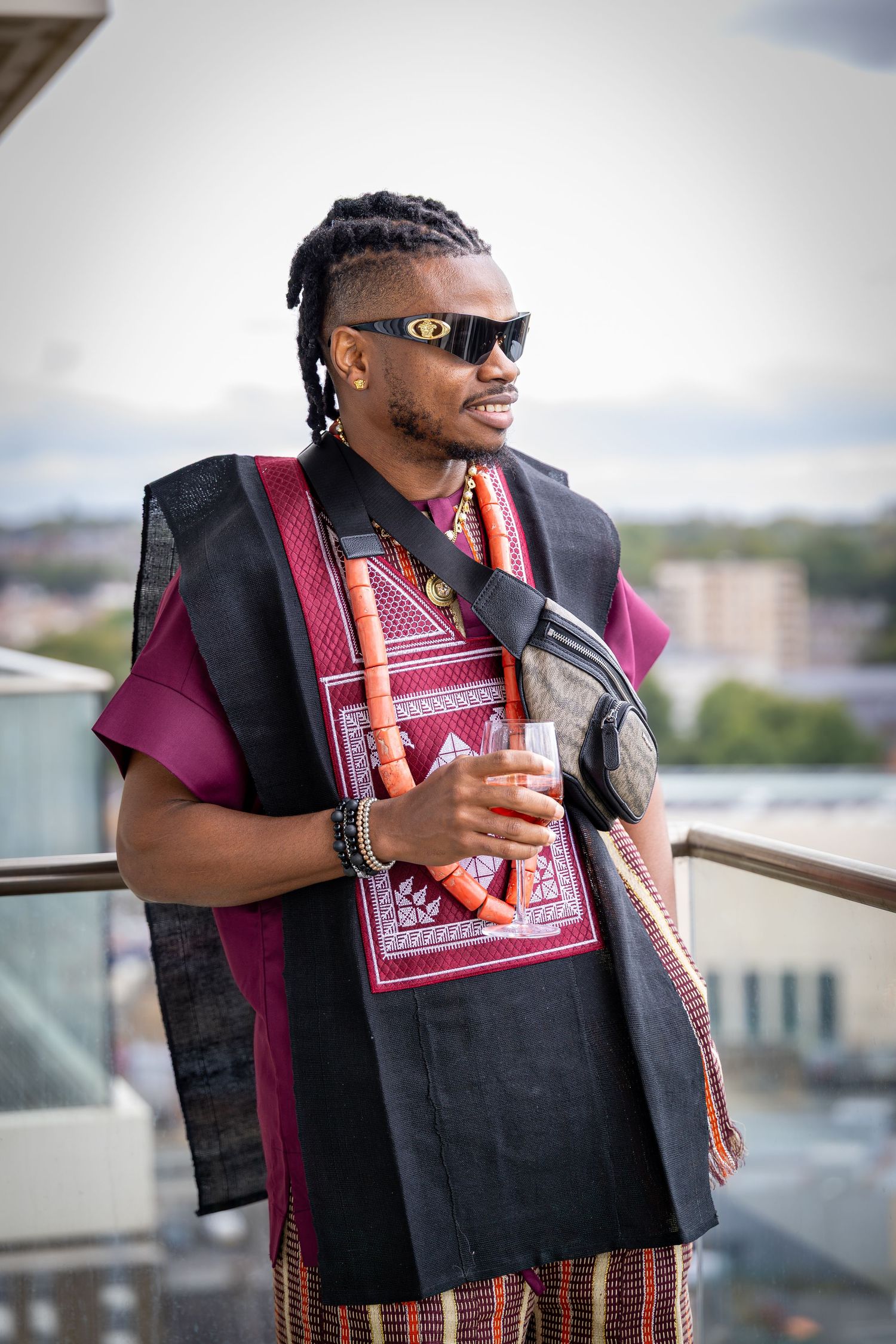 Man in traditional African attire with coral beads and sunglasses holding a drink on a rooftop balcony.