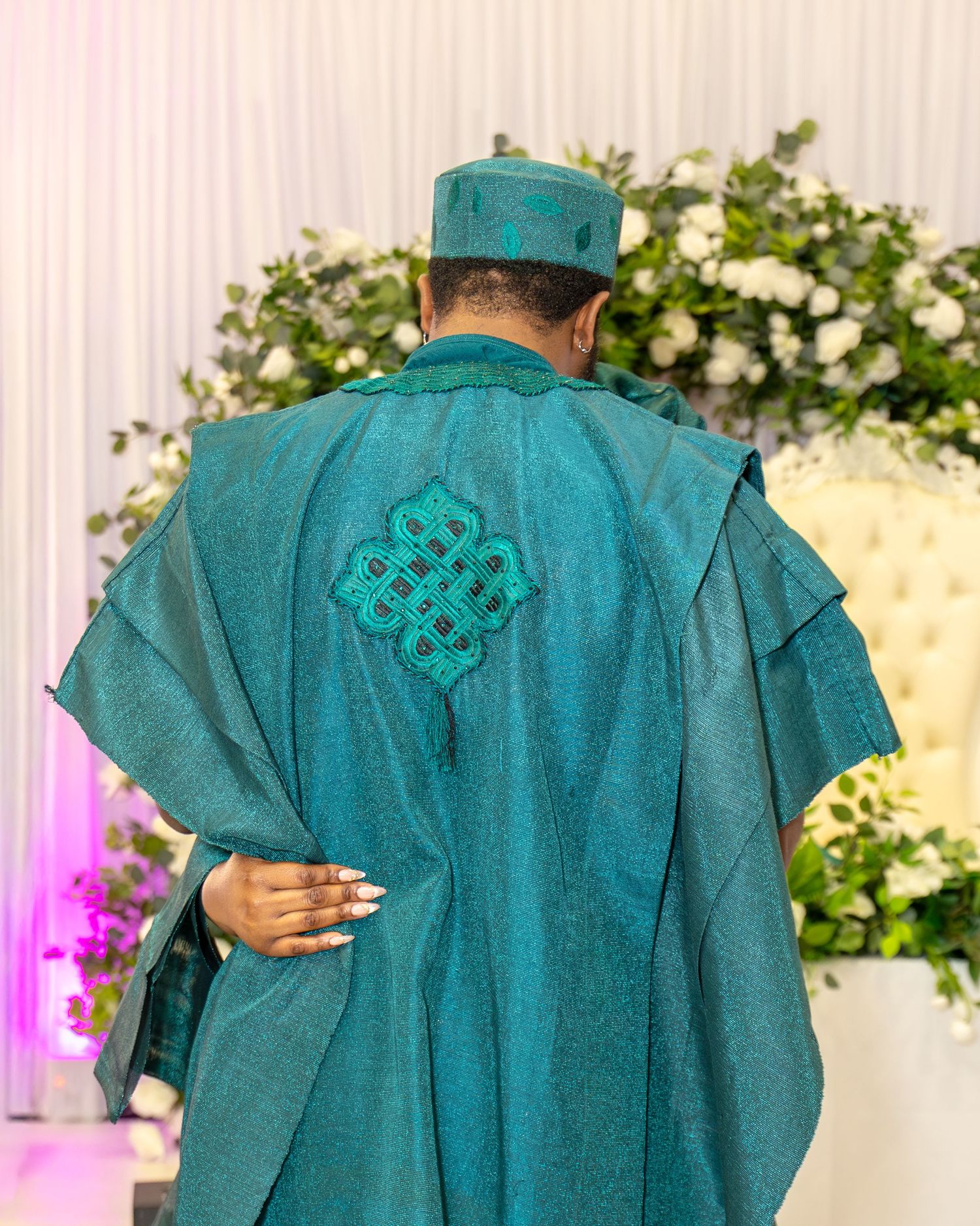 A man in a teal Nigerian agbada with intricate embroidery and matching cap stands before a floral backdrop.