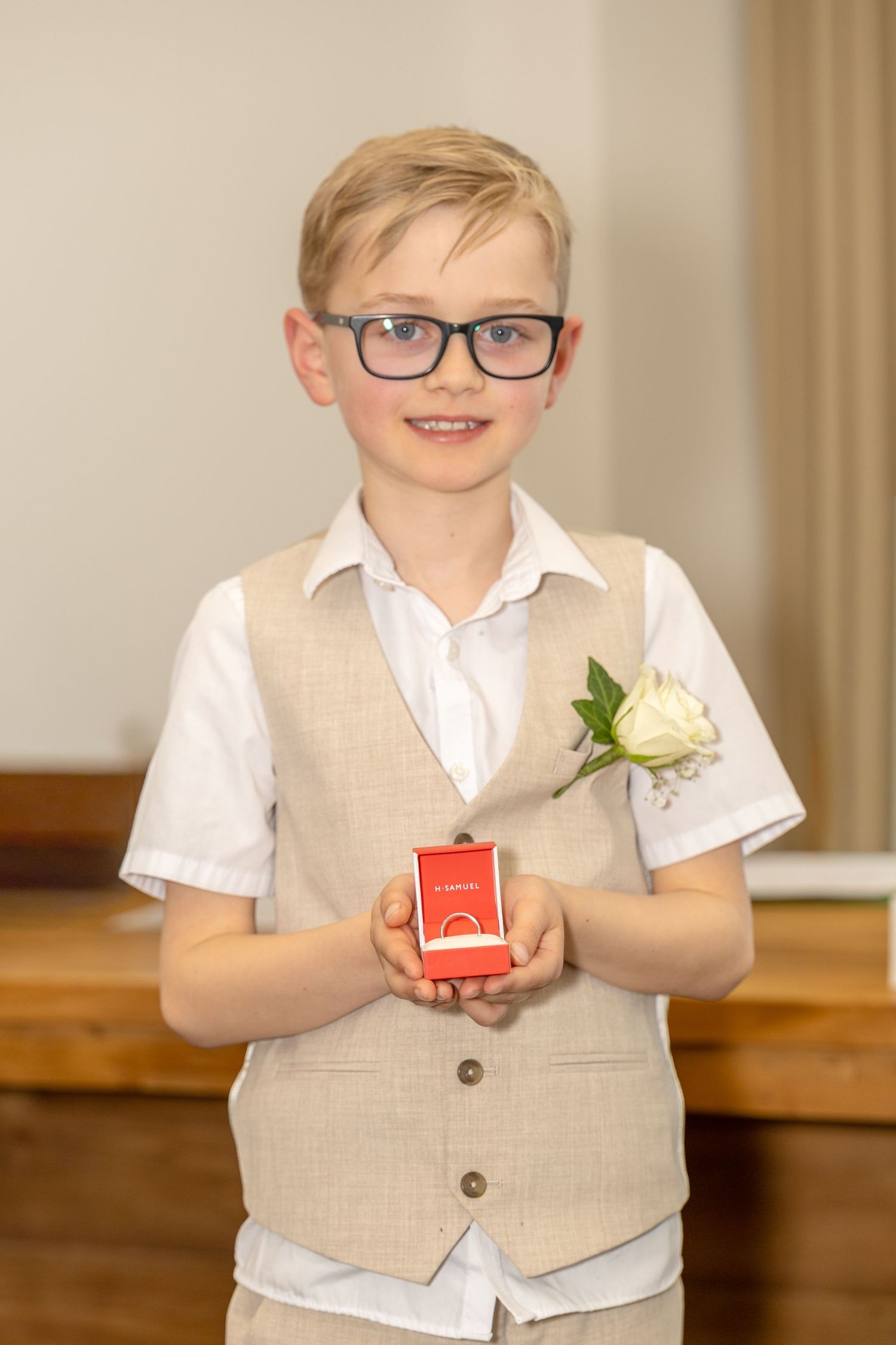 Young ring bearer in beige vest holding a red jewelry box at a wedding ceremony.