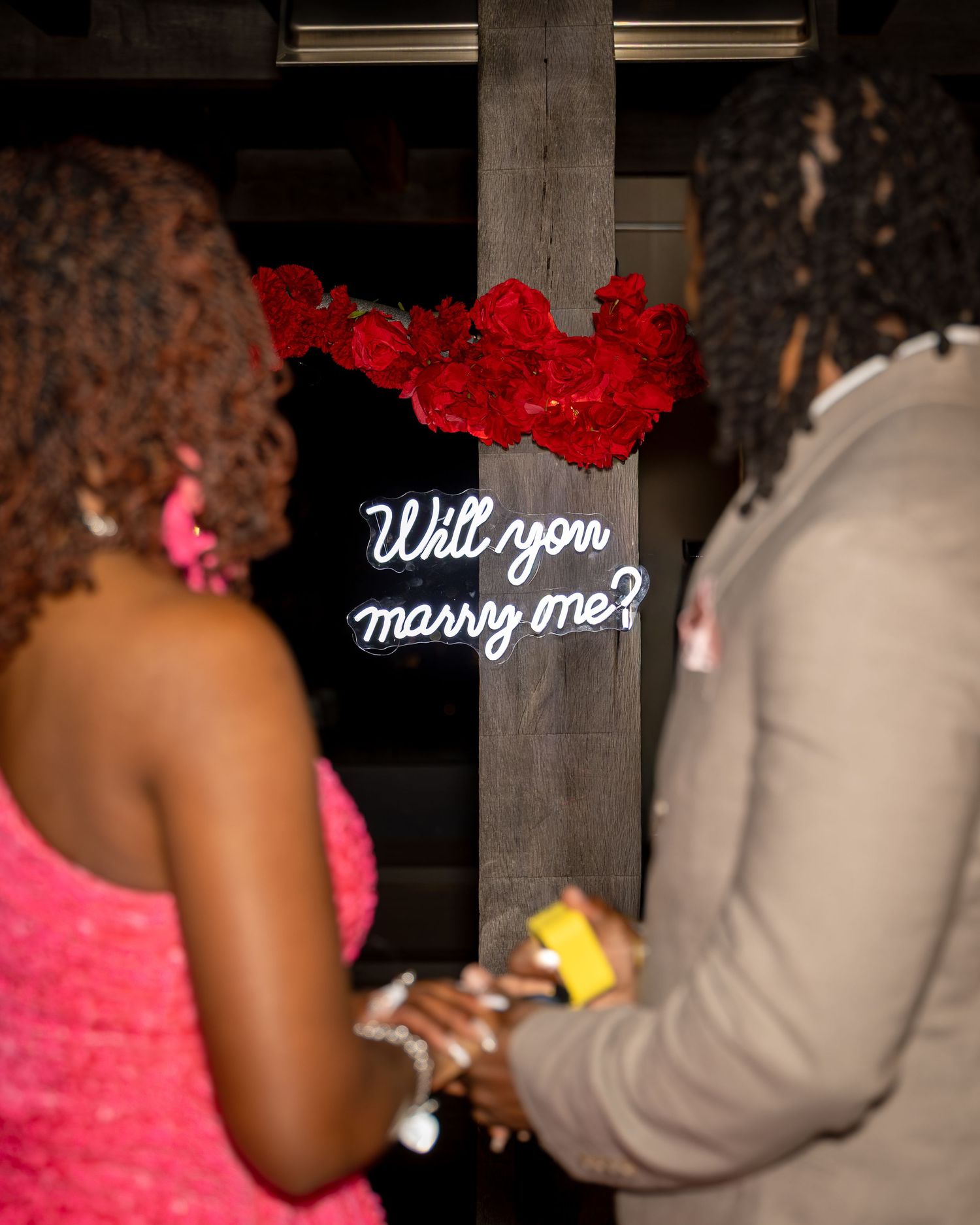 Couple holds hands near Will You Marry Me neon sign surrounded by red roses at night.
