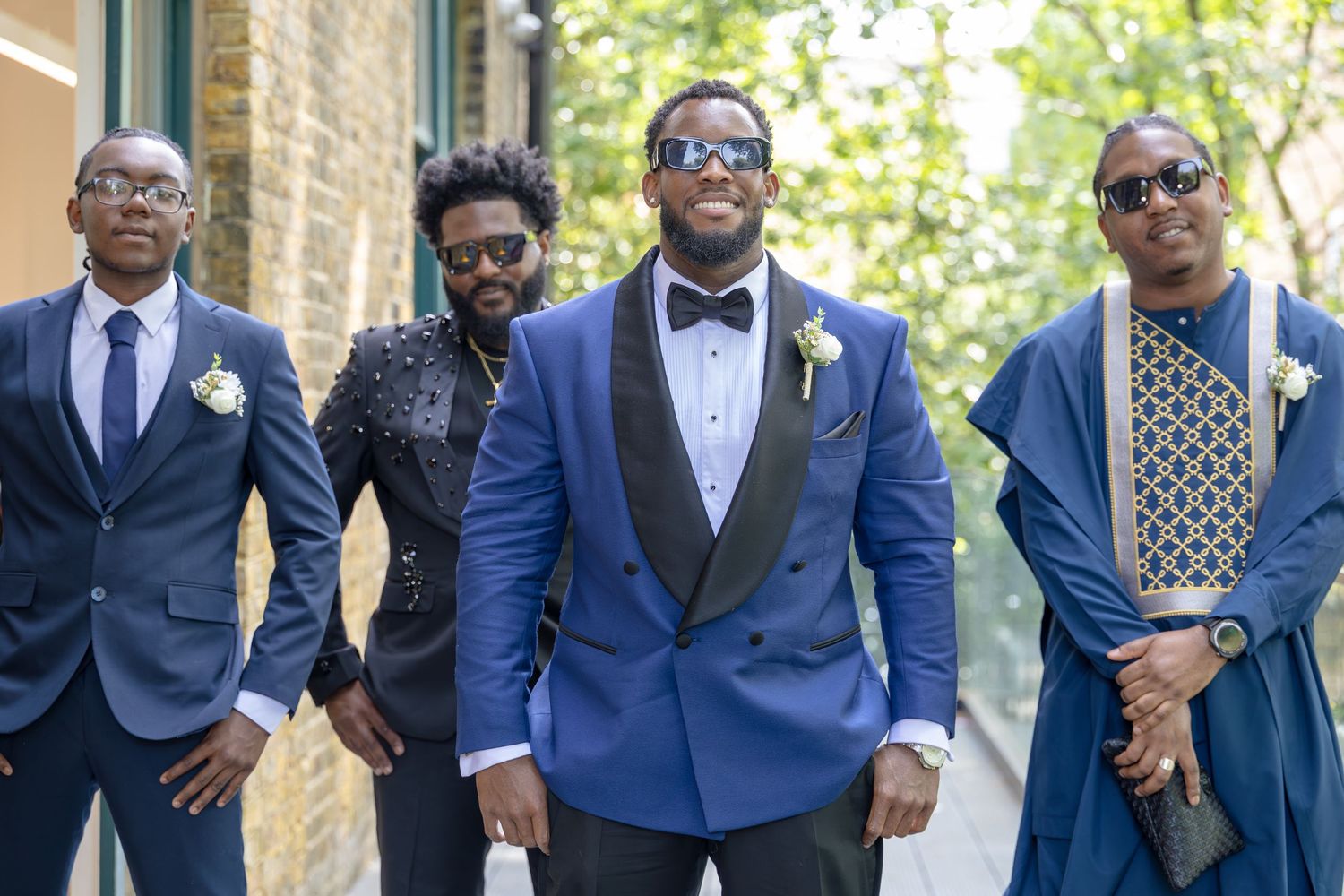 Four well-dressed Black men in formal suits and bowties pose outdoors, with the groom in a royal blue tuxedo centerpiece.
