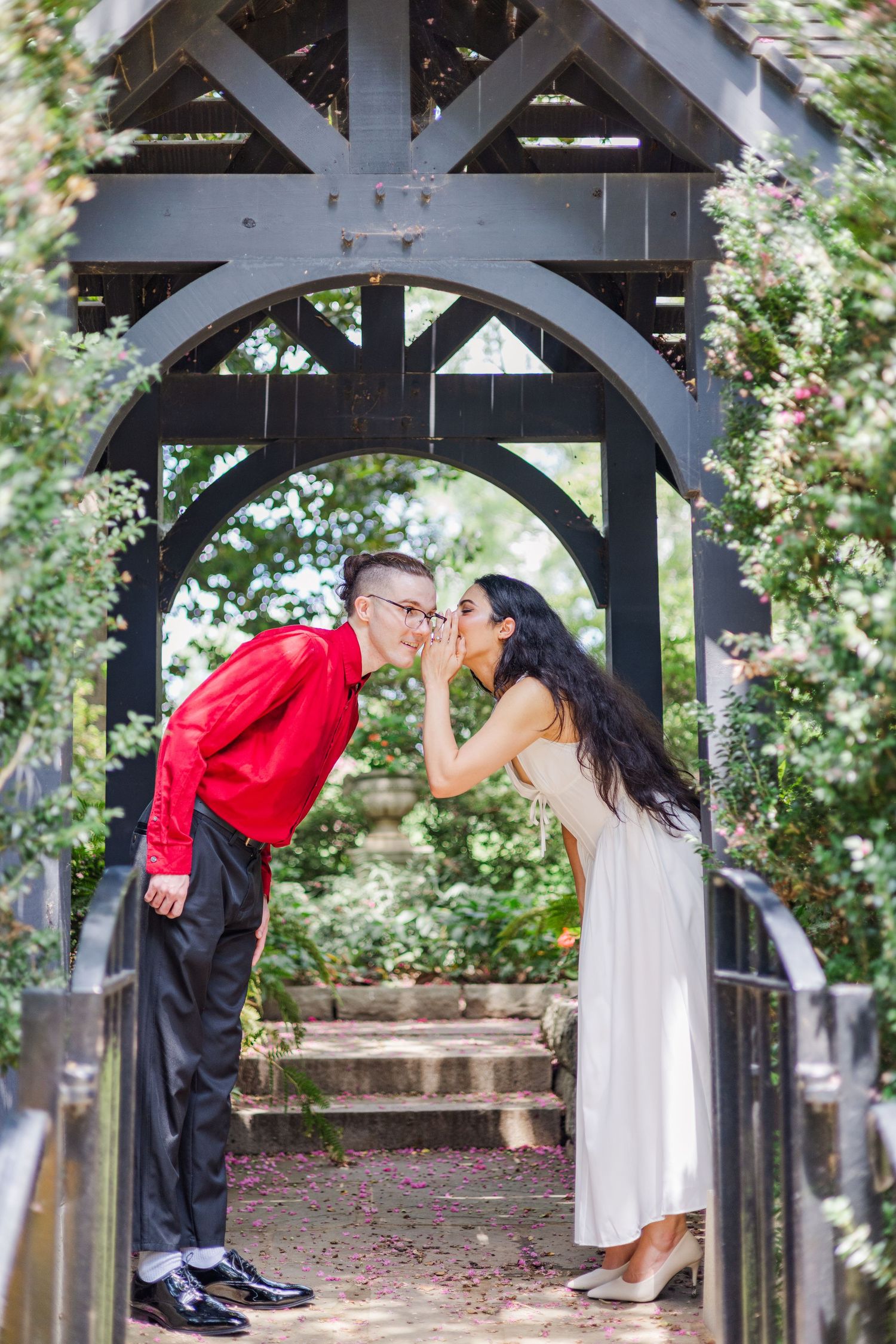 A couple shares a playful kiss under a wooden garden archway surrounded by greenery.
